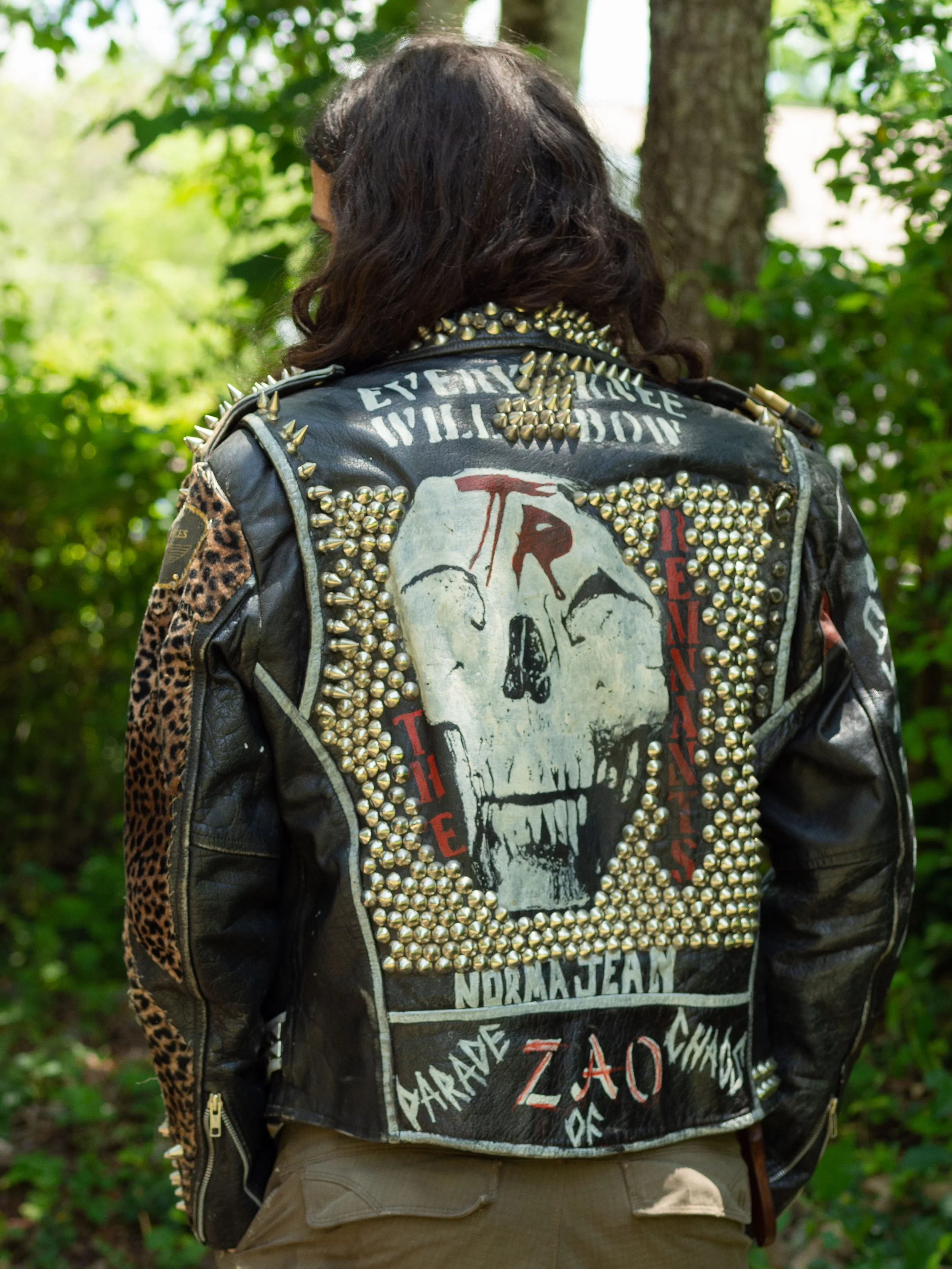 Person wearing a studded leather jacket with skull design and text, standing outdoors in a wooded area.
