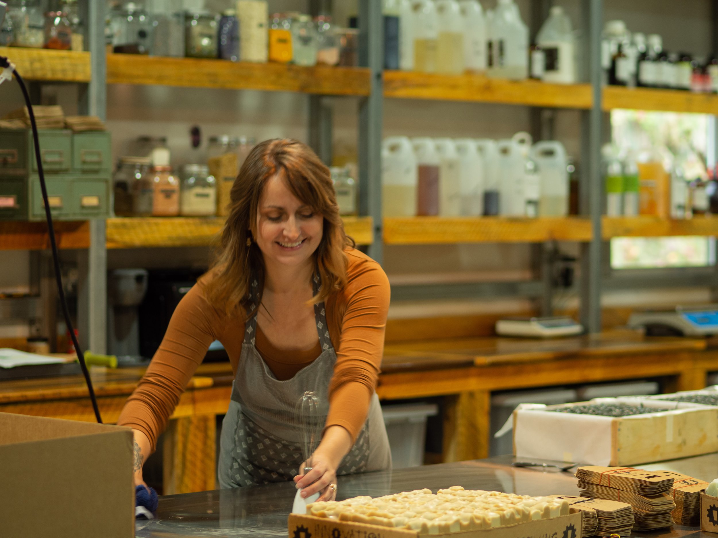 Woman cleaning a table in soap making workshop with shelves of ingredients in background.