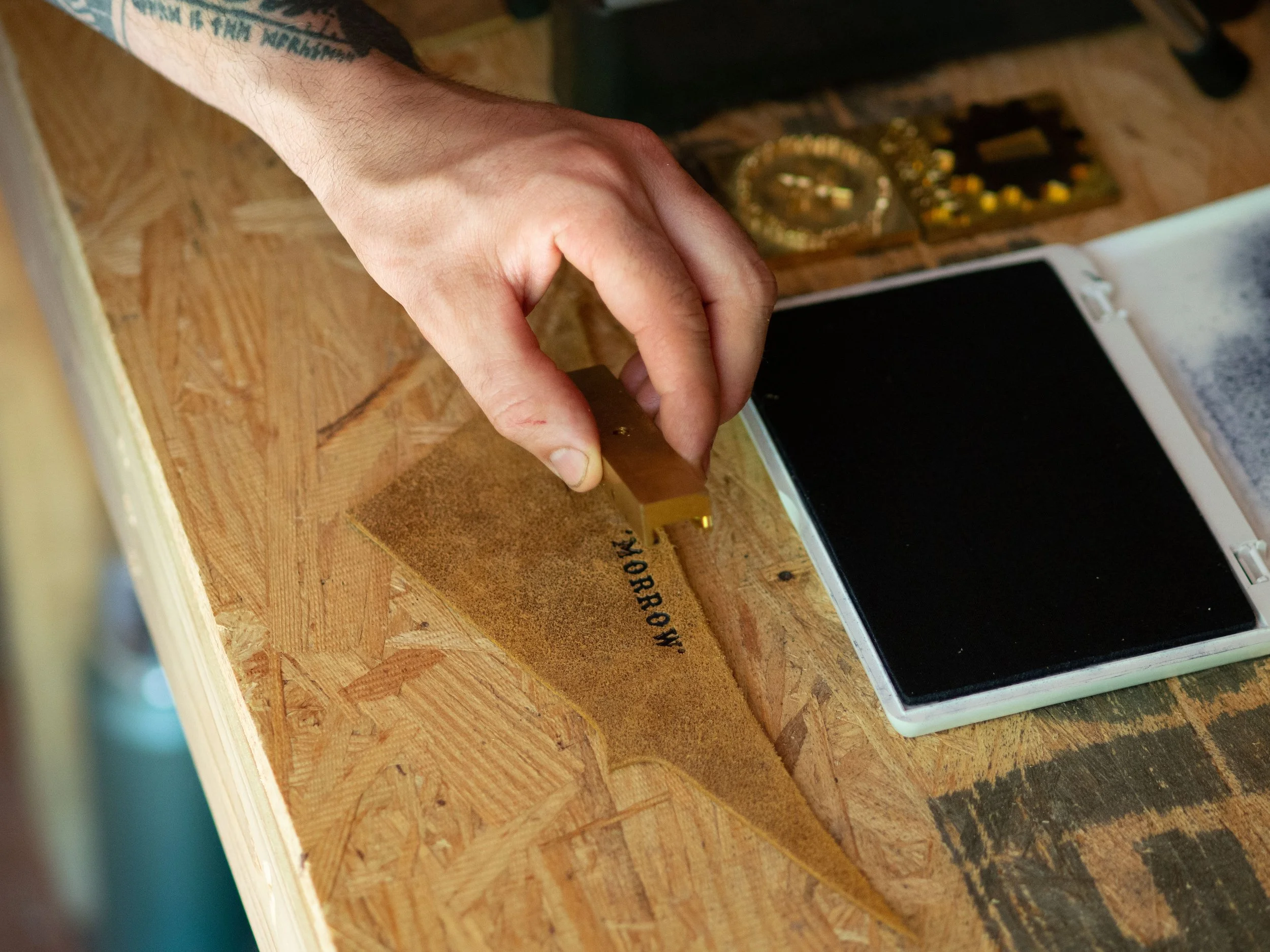 Person holding a leather piece with 'TOMORROW' stamped on it, next to an ink pad and gold stamping tools on a wooden surface.