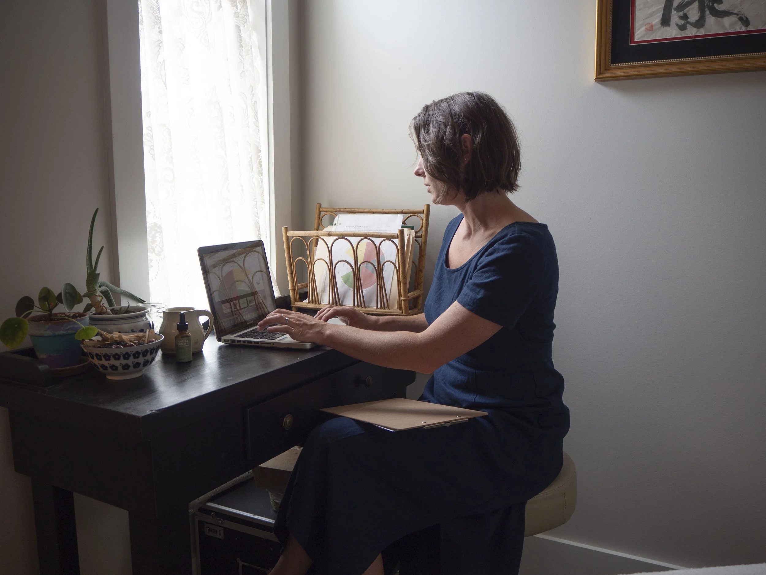 Woman in blue dress working on laptop at a desk by a window with plants and decor