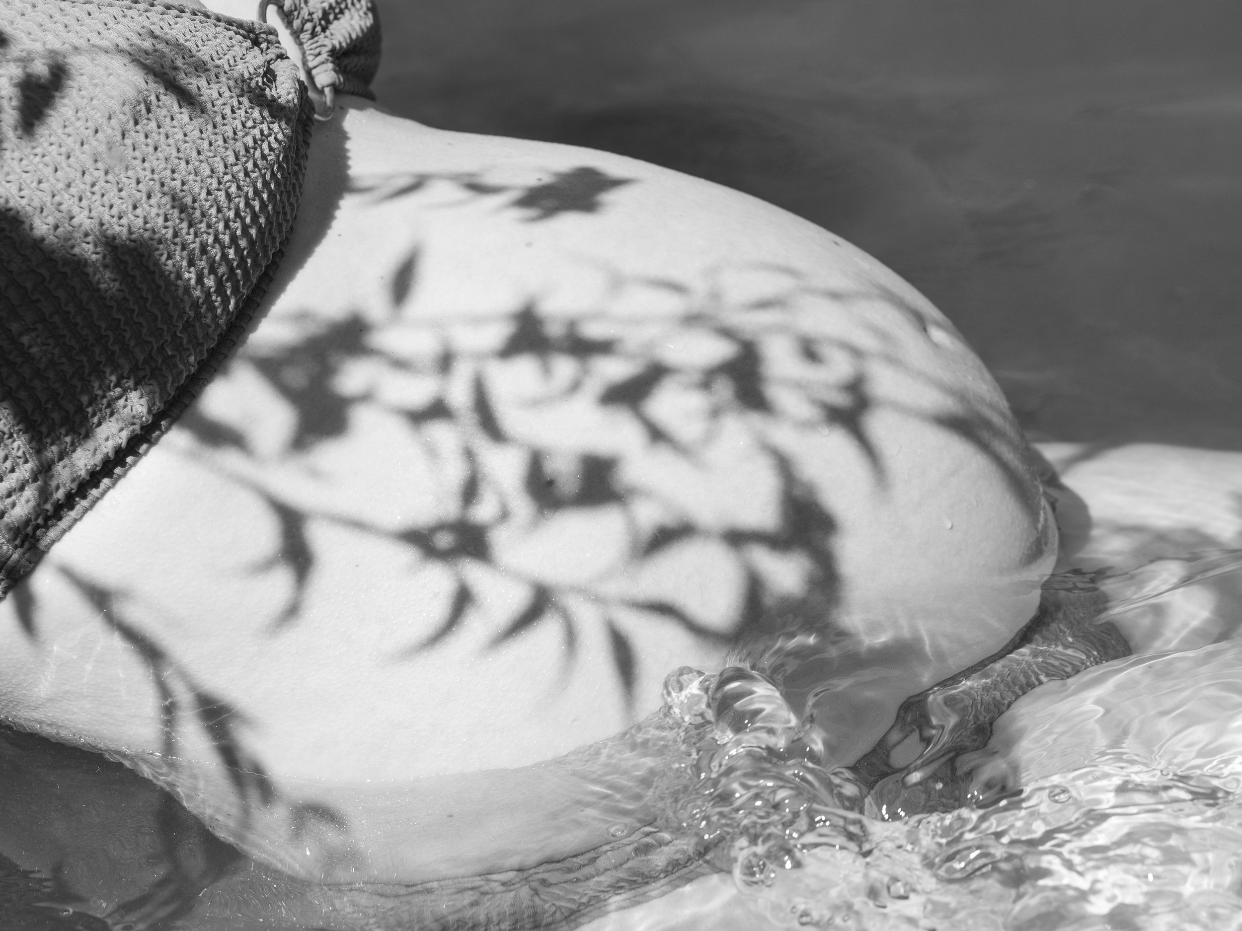 Black and white photo of a pregnant belly with leaf shadows and water rippling against it.