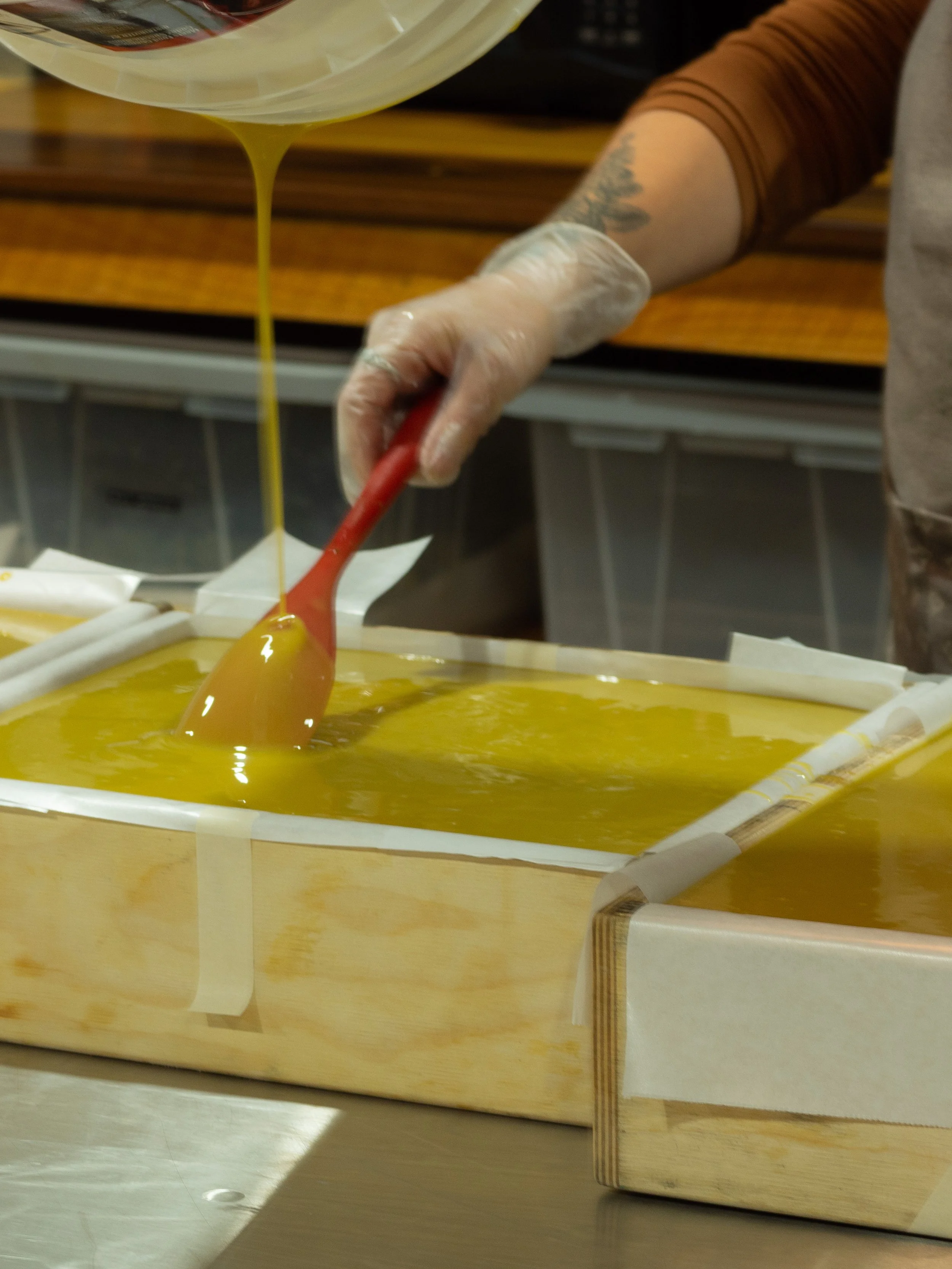 Person pouring yellow liquid soap mixture into a mold, using a spatula to spread it.