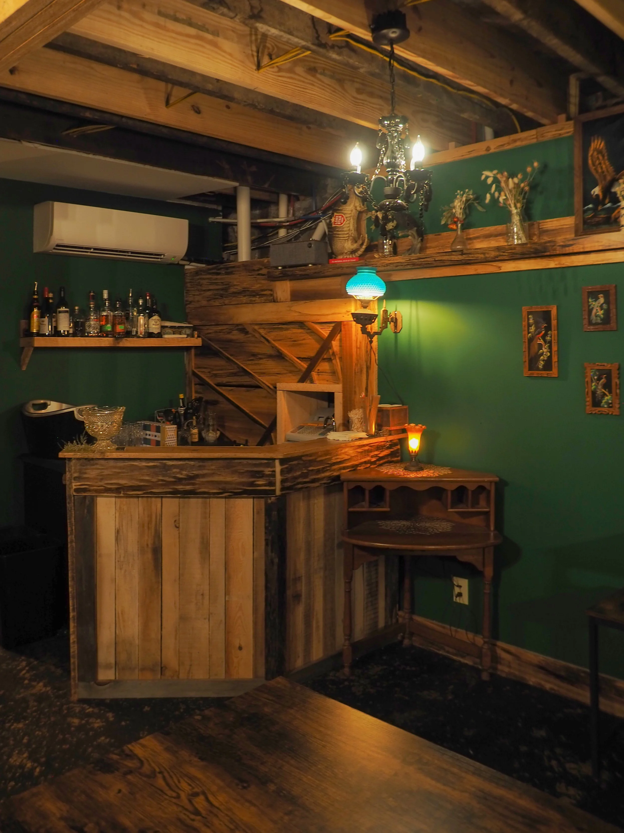 Cozy home bar with wooden counter, green walls, bottles on shelf, vintage chandelier, and decorative items.