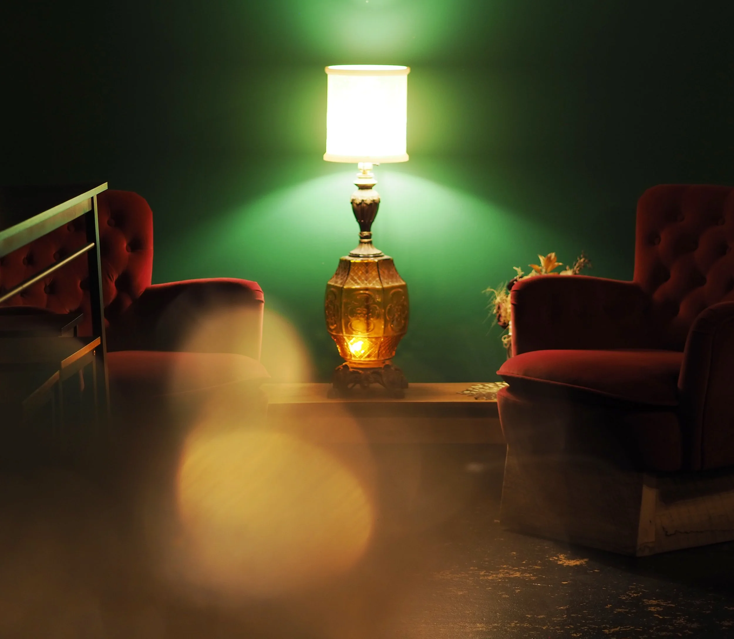 A dimly lit room featuring a vintage-style lamp on a wooden table between two burgundy armchairs against a green wall.