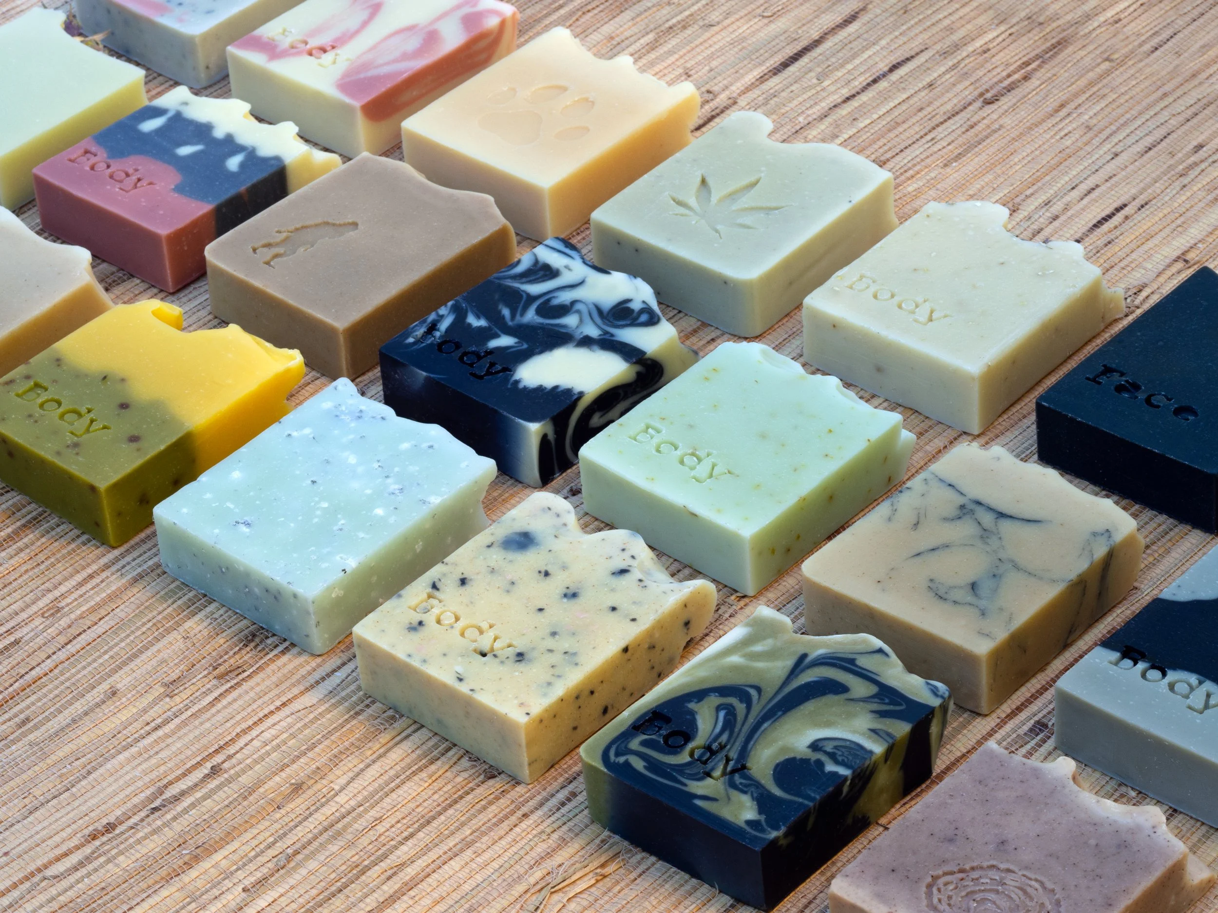 Assorted colorful handmade soap bars arranged neatly on a wooden surface, some engraved with the words 'body' and 'face' and featuring decorative patterns and emblems.