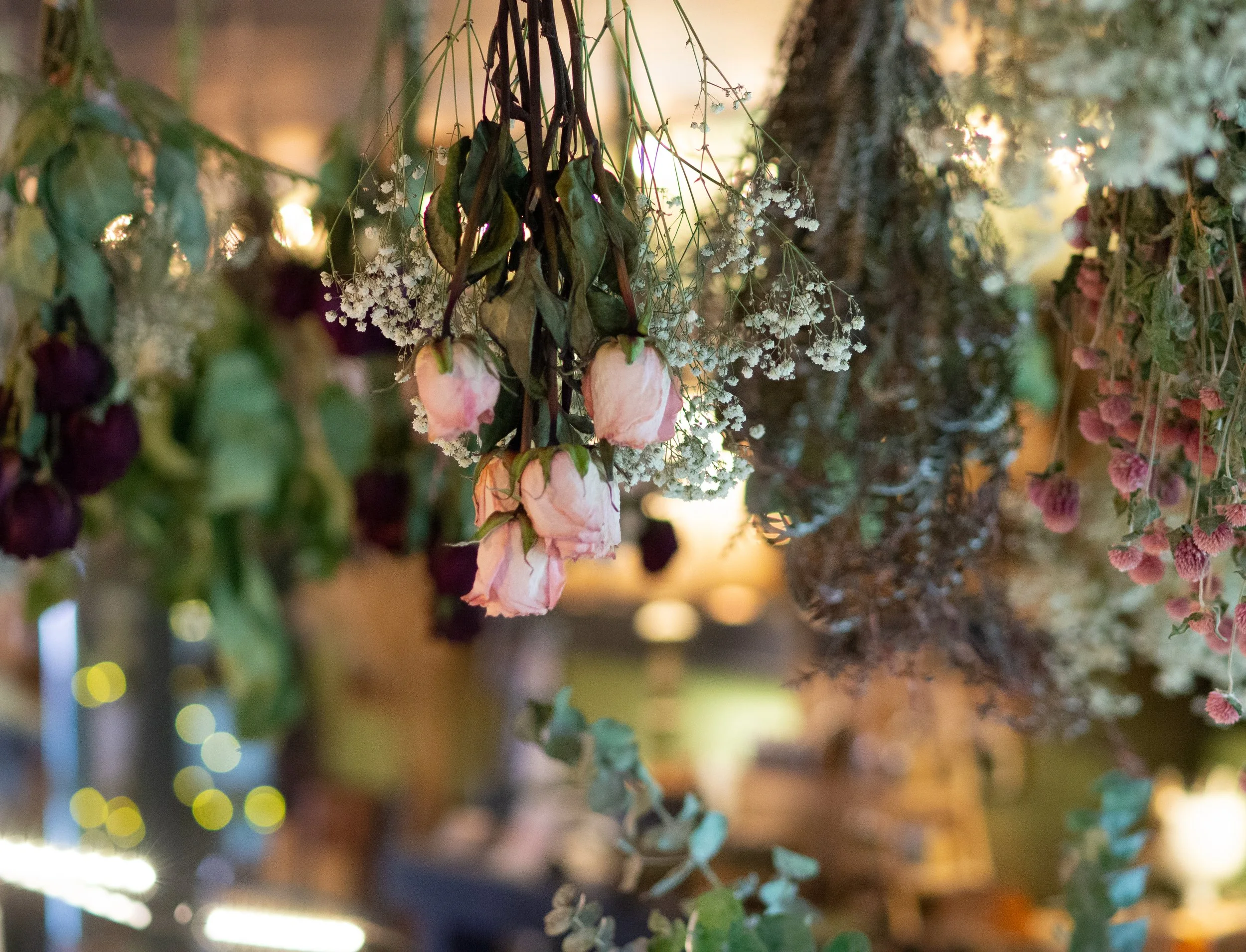 Dried flowers hanging upside down, including pink roses, baby's breath, and other assorted foliage, with a blurred background.