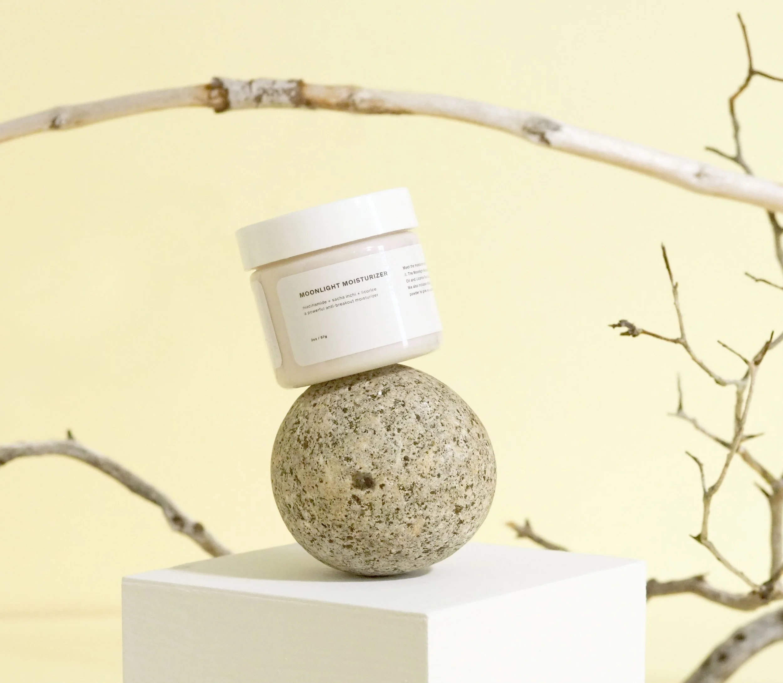 A jar of Moonlight Moisturizer balanced on a rough stone sphere, placed atop a white pedestal against a beige background with bare branches.