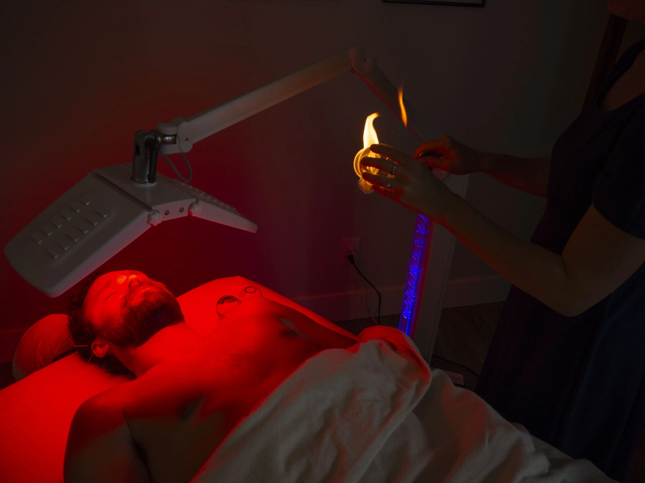 A person lying under a red light therapy lamp, with another individual holding a fire cupping tool.