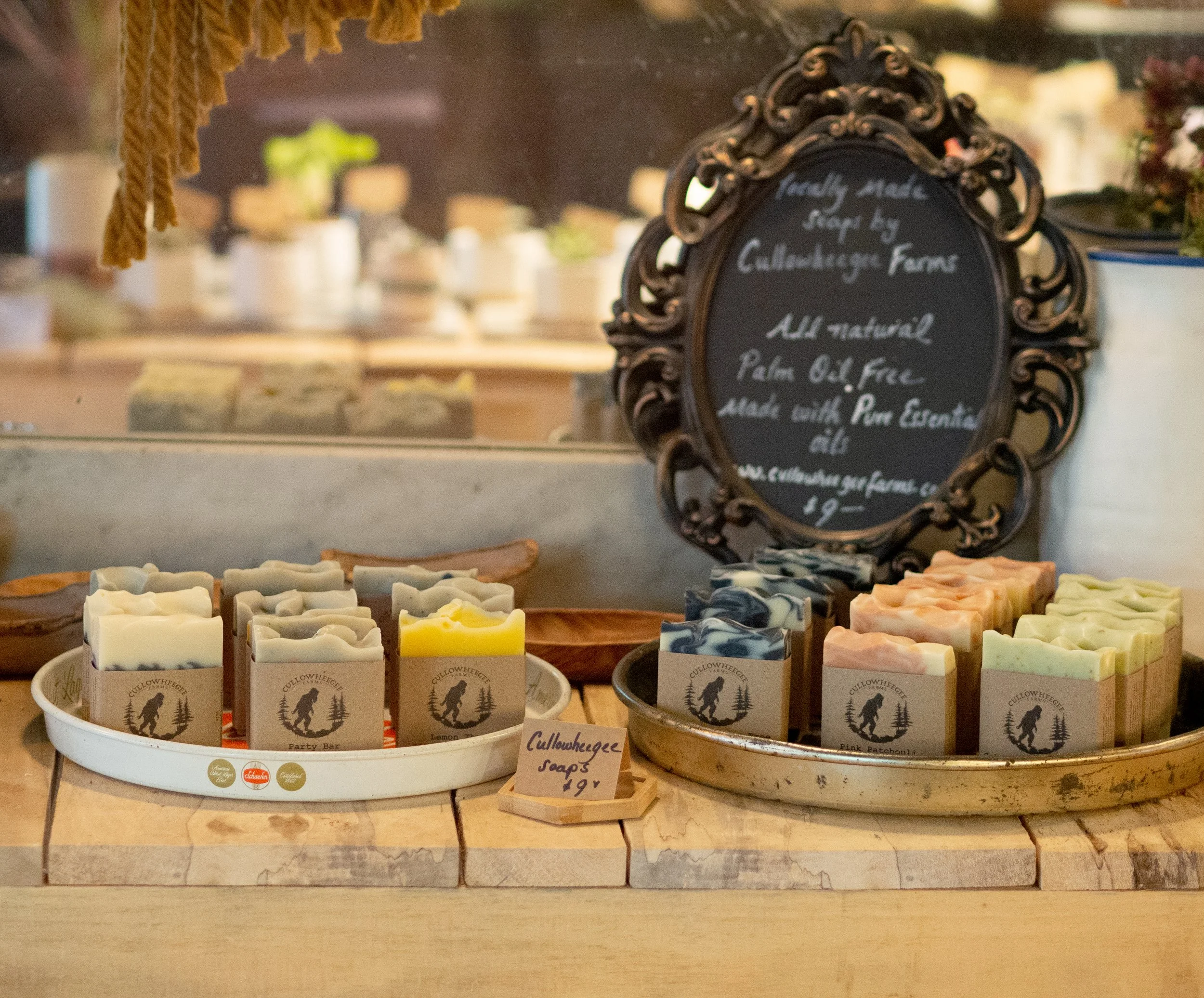 Display of handcrafted soaps on a wooden table, with soap bars arranged on white and gold trays. A decorative chalkboard sign behind them reads "freshly made soaps by Cullowheegee Farms, all natural, palm oil free, made with pure essential oils." A s