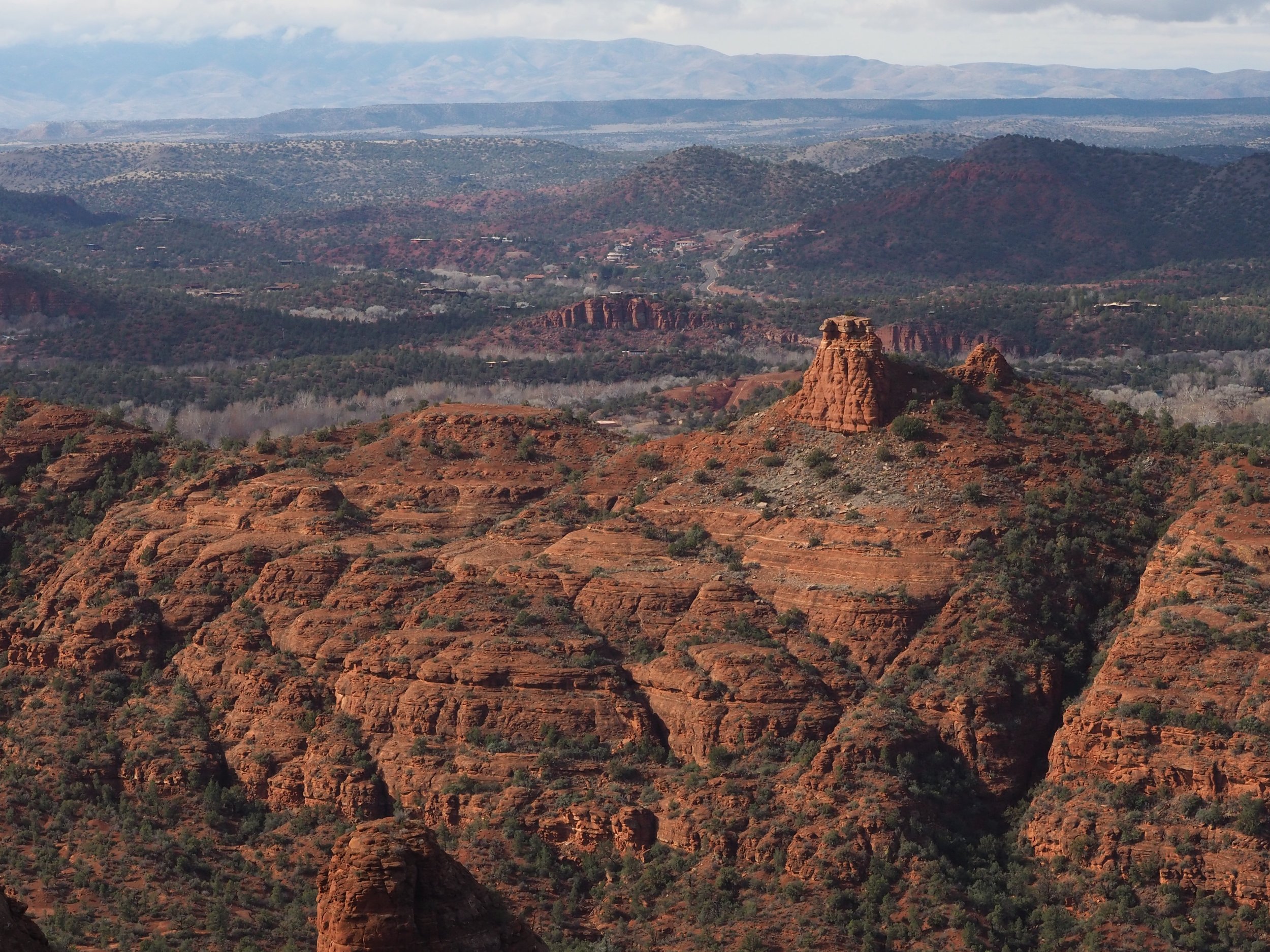 Landscape view of red rock formations and mesas in Sedona, Arizona, with scattered vegetation and distant mountains under a partly cloudy sky.