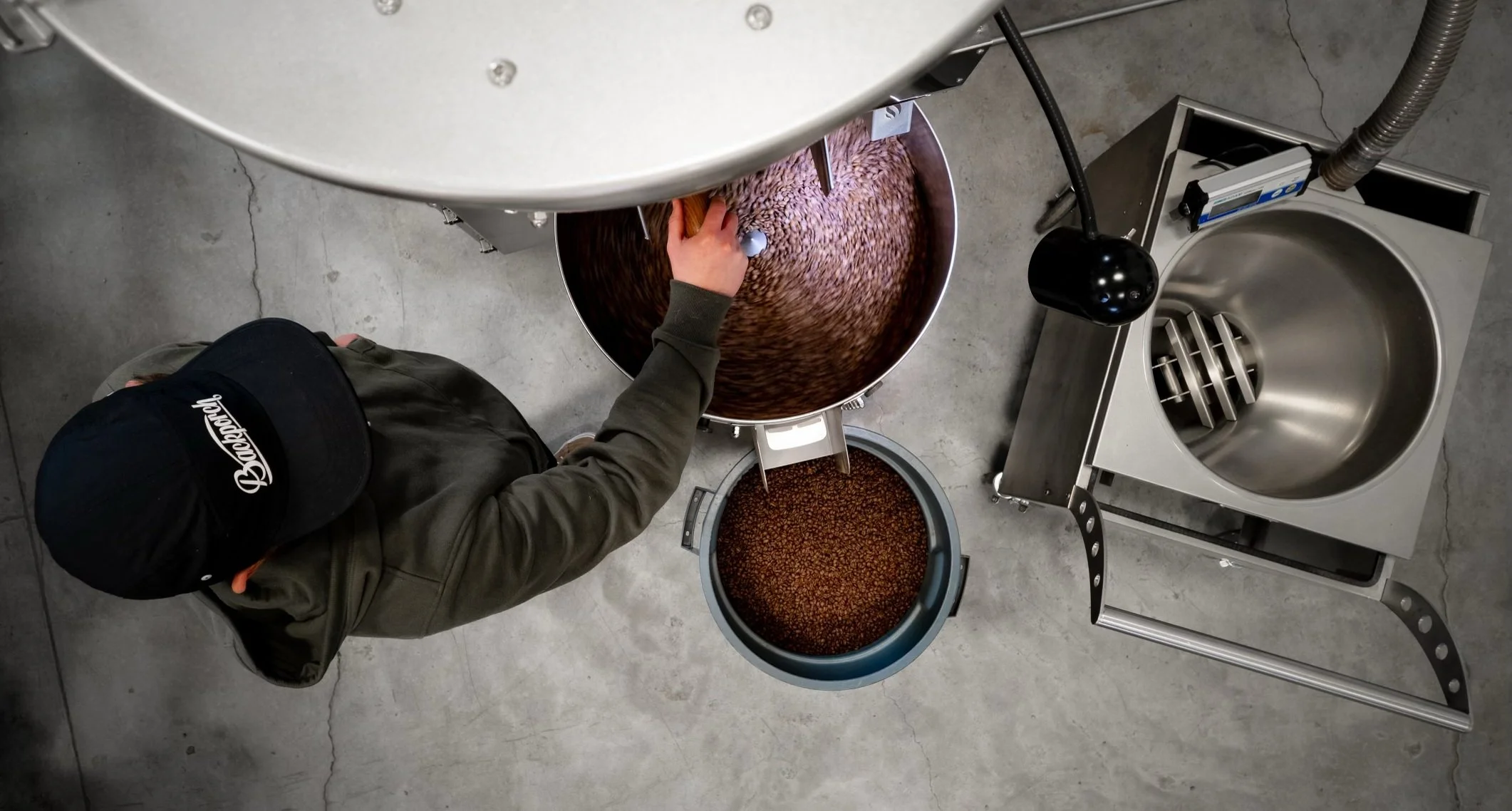 Bird's-eye view of a person in a cap operating a coffee roasting machine. The roaster is filled with roasted coffee beans, and another container holds more beans. Nearby, there's a shiny metal bowl.