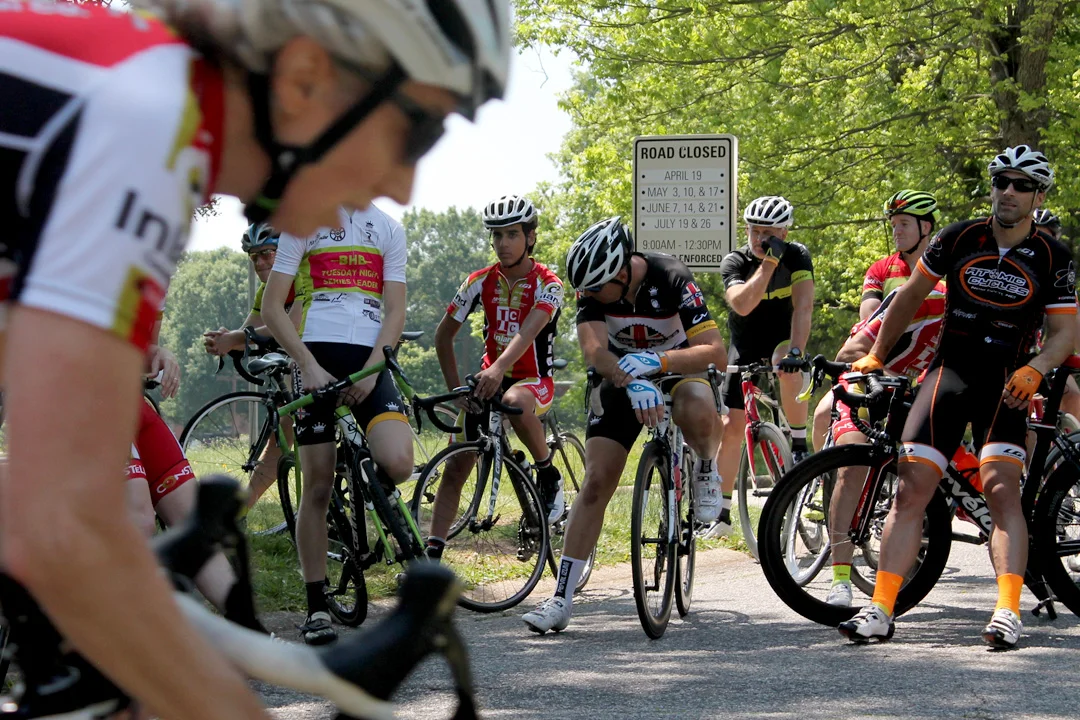 Waiting for their race to begin, riders watch the women's event. A street sign tells motorists about road closure dates. In the foreground is Robin Baxter.