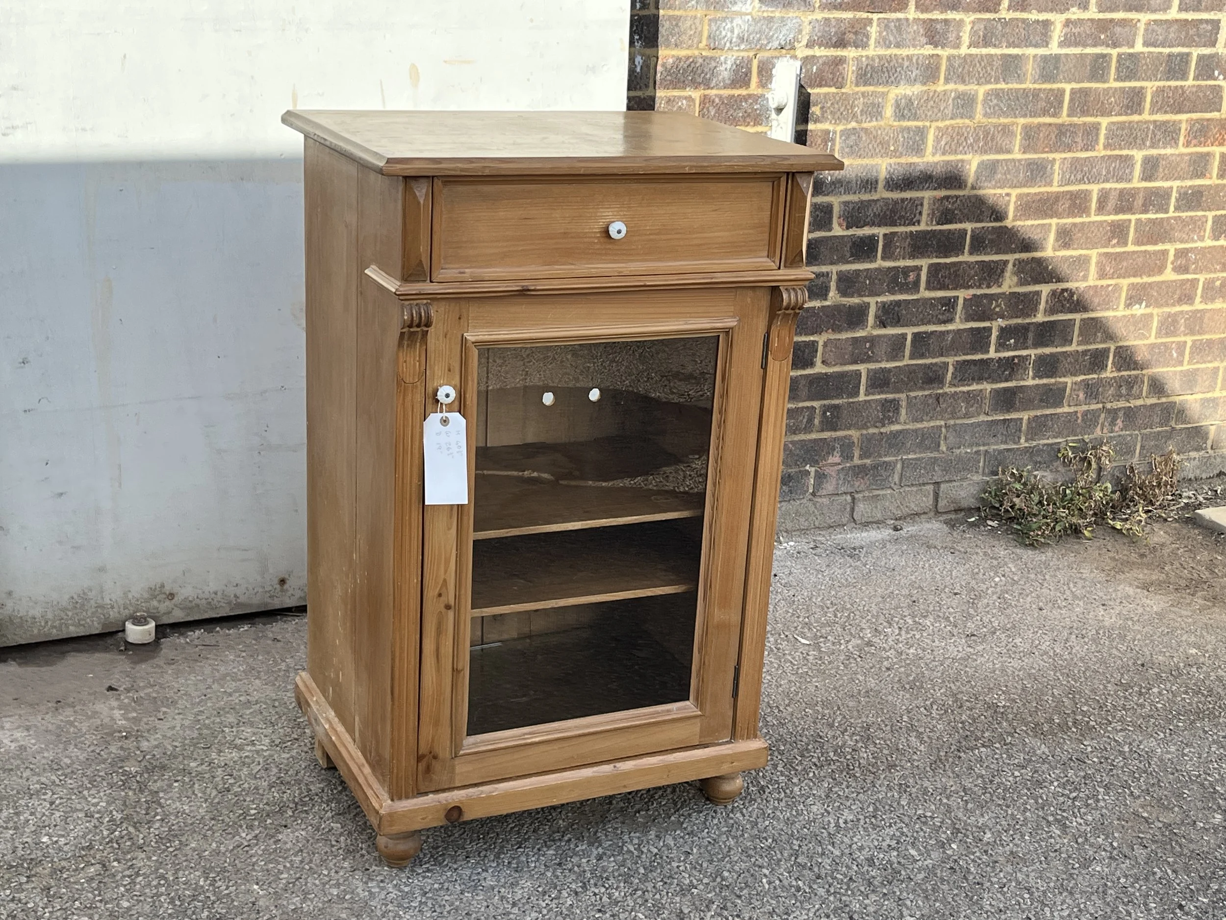 Angled view of the small pine cupboard with a glazed door, internal shelves and upper drawer