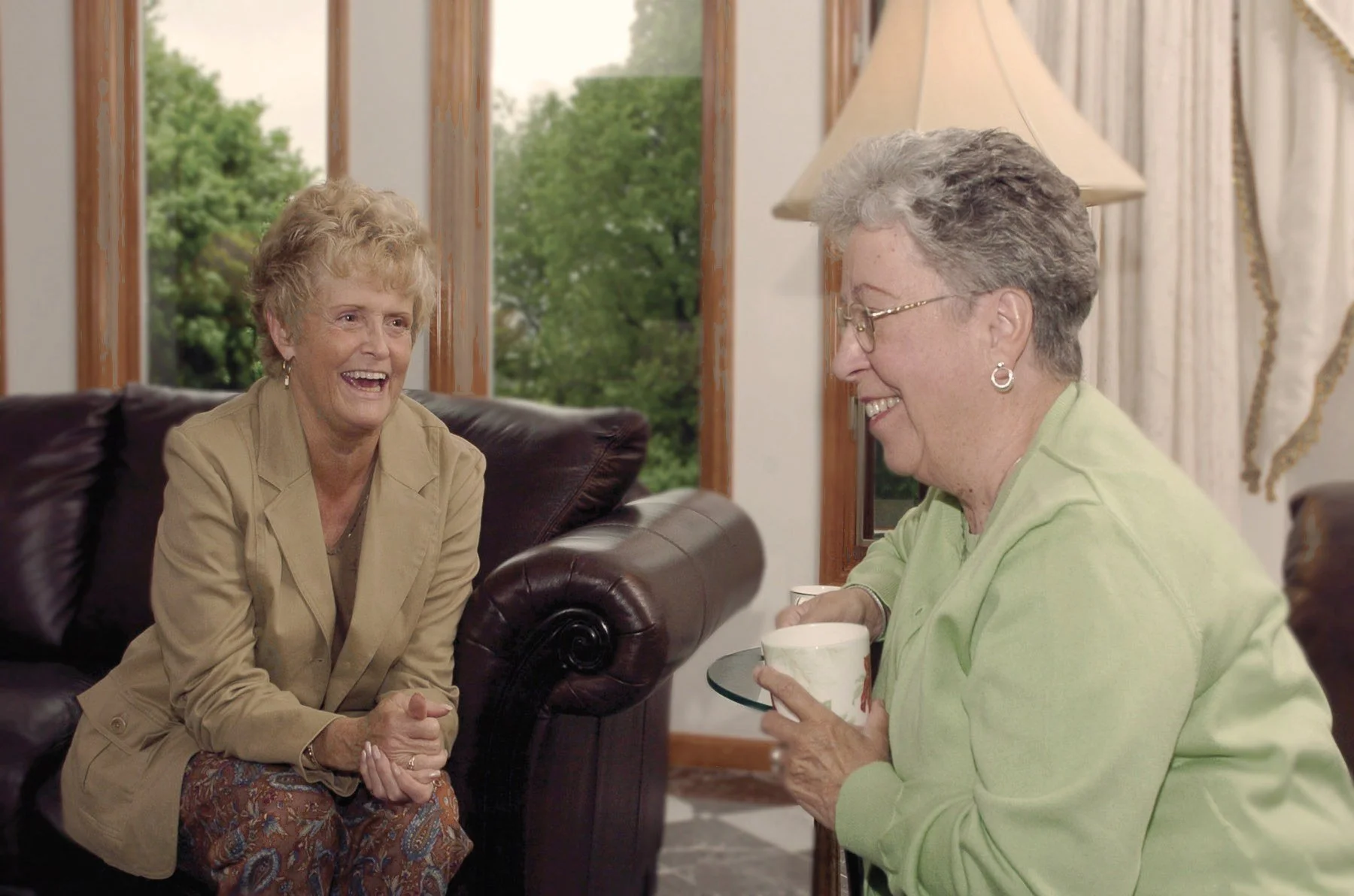 Two older women laughing together over a cup of tea in a sitting room.