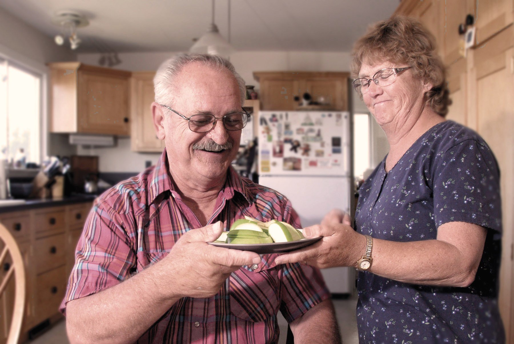 A carer hands an older man a plate of sliced apple in his kitchen.