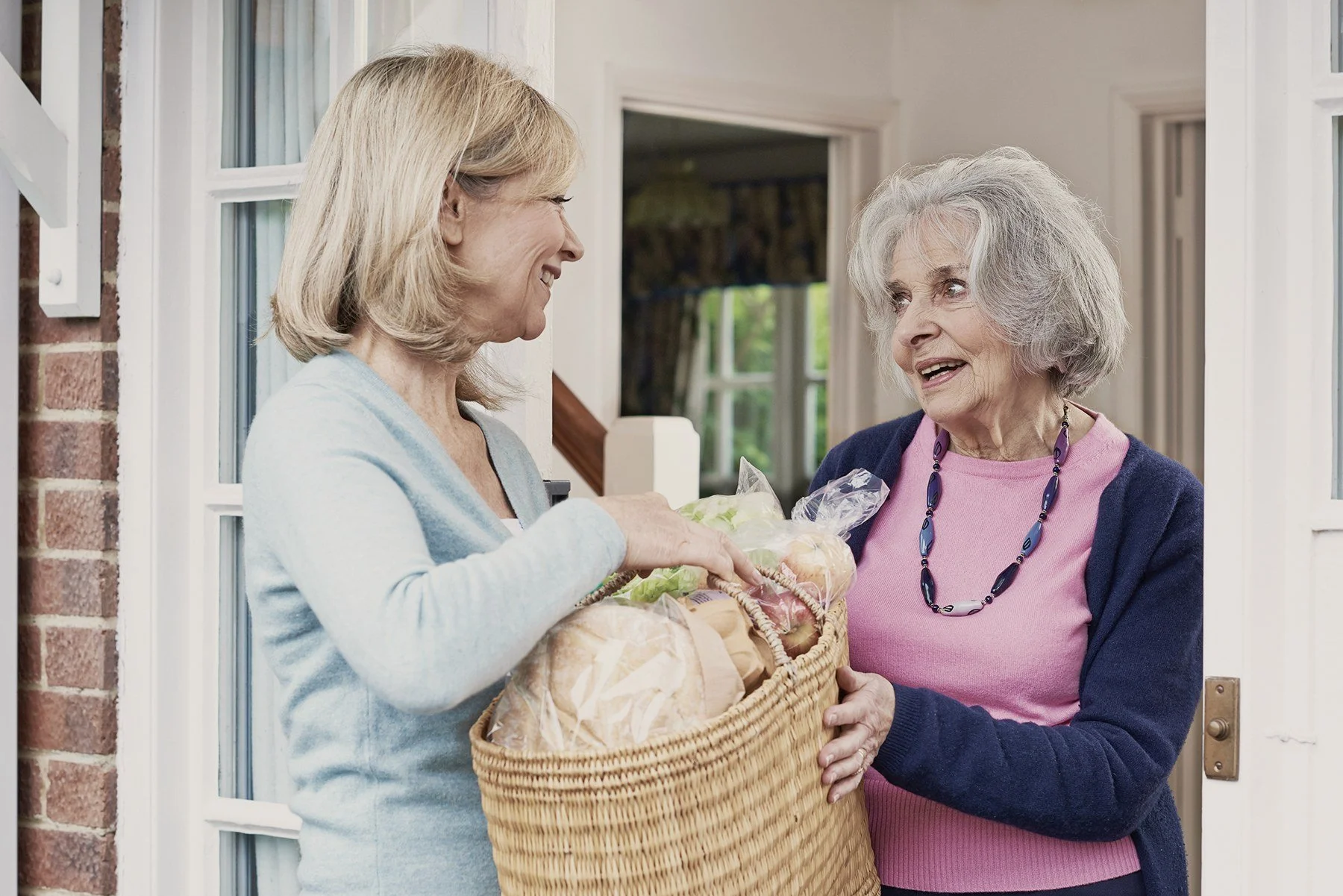 A woman arrives at an older friend's front door with a basket of shopping, and they greet each other warmly.