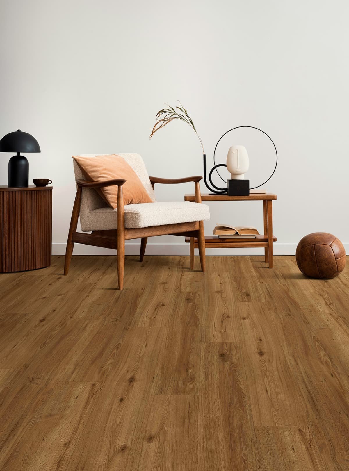 Minimalist living room with a beige armchair, orange cushion, black side table with black lamp, wooden side table with decorative objects including a white vase, dried plant, and book, and a brown leather pouf on hardwood floor against a plain white wall.