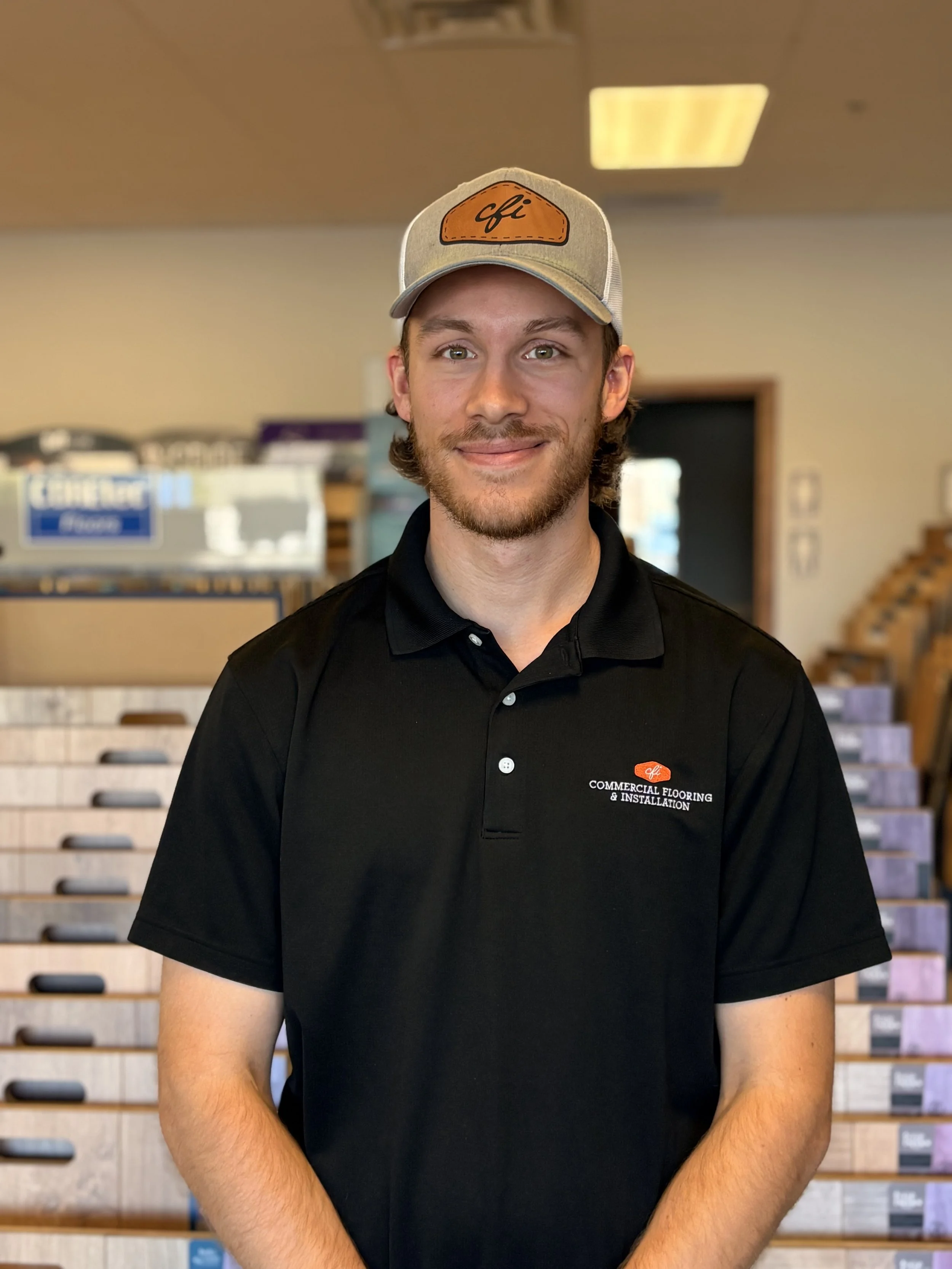 A young man with light brown hair, a beard, and green eyes wearing a beige baseball cap with an orange patch labeled 'cfi' and a black polo shirt with a logo reading 'Commercial Flooring & Installation'. He is smiling and standing indoors with a background of shelves and boxes.