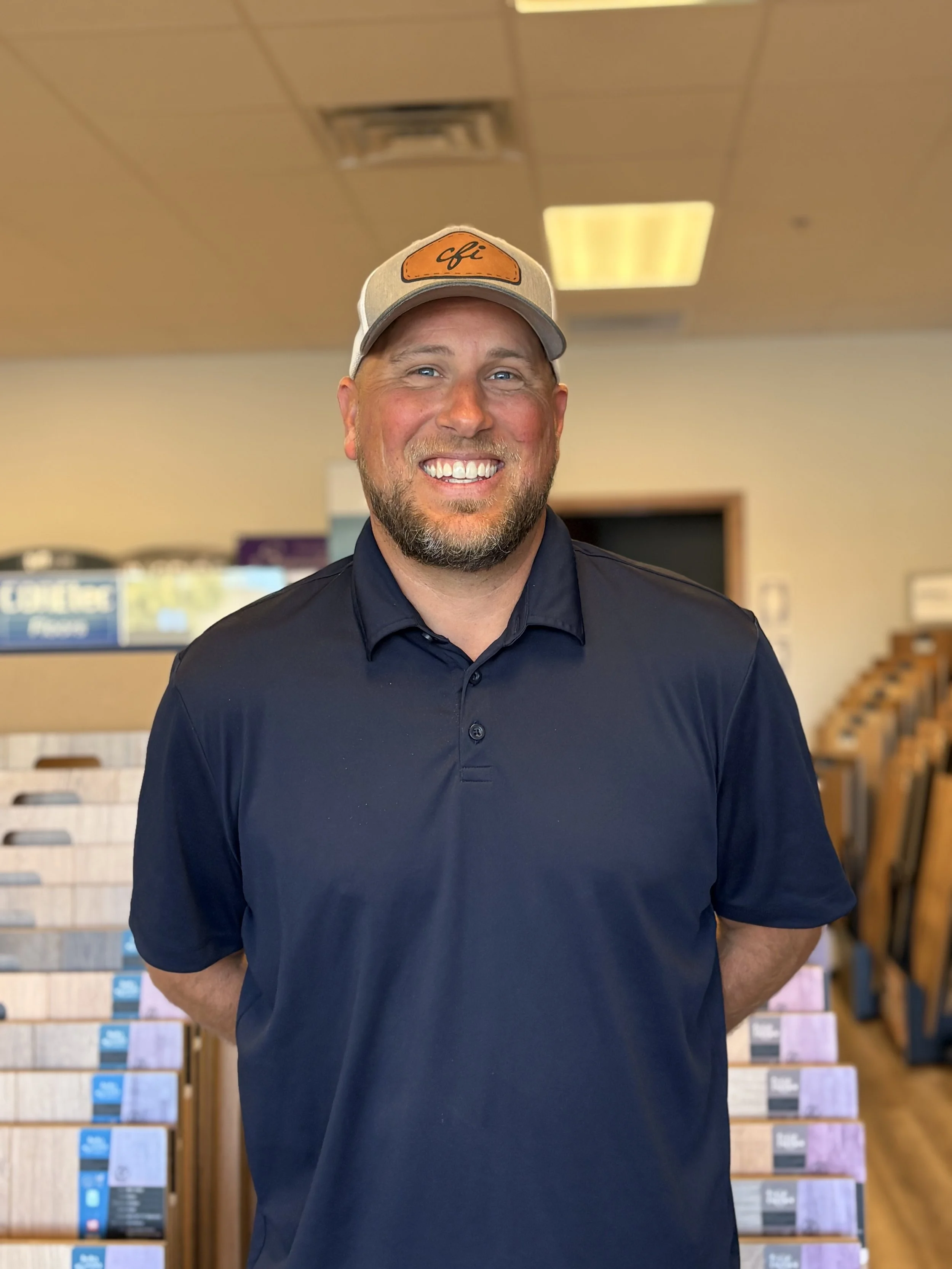 A smiling man with a beard wearing a gray cap with orange patch and the word 'cfi' on it, and a dark navy polo shirt, standing in a room with wooden shelves and various products or samples behind him.