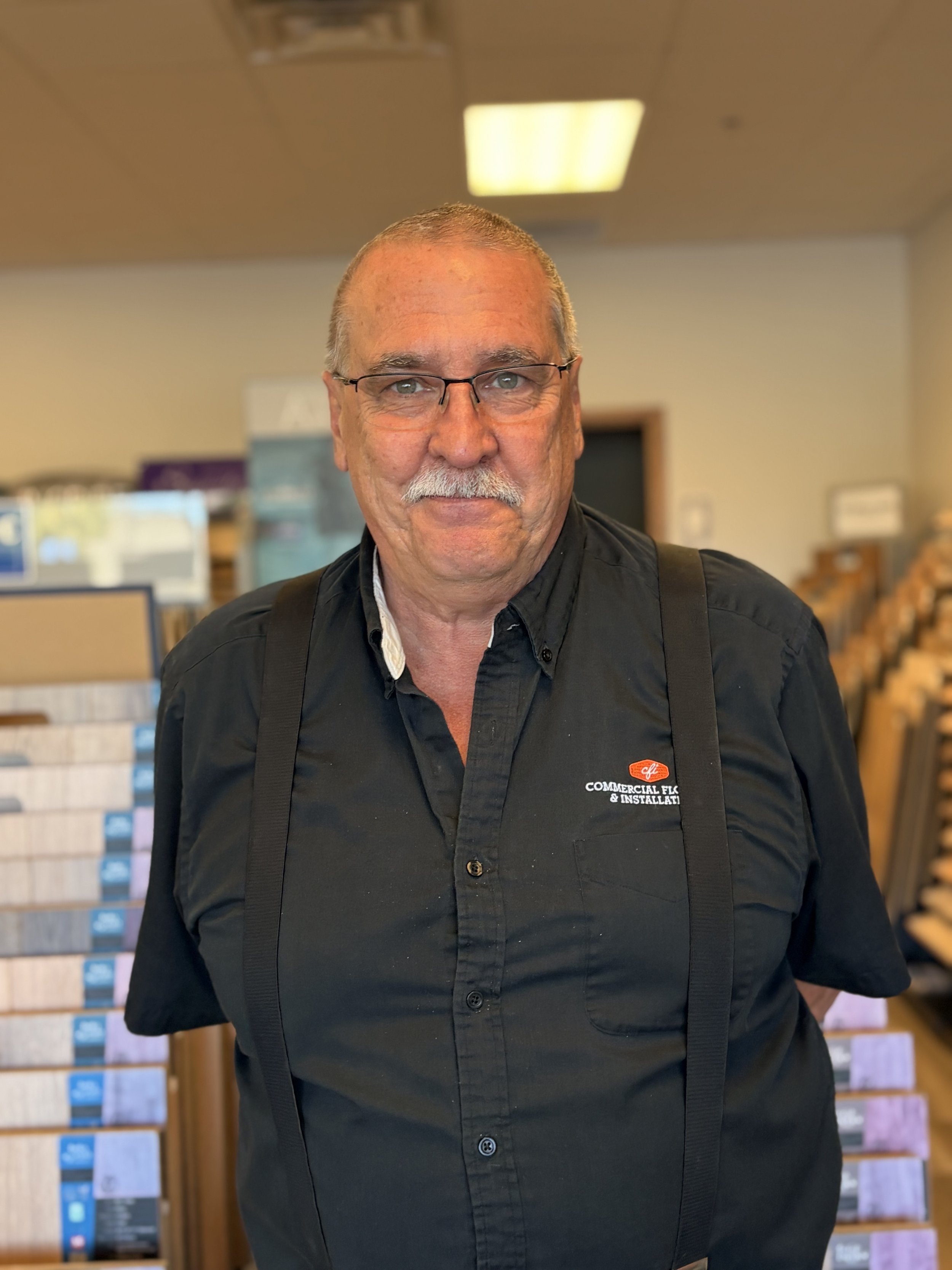 A middle-aged man with glasses and a mustache smiling in a hardware or home improvement store. Behind him are display boards with samples of tiles or wall coverings.