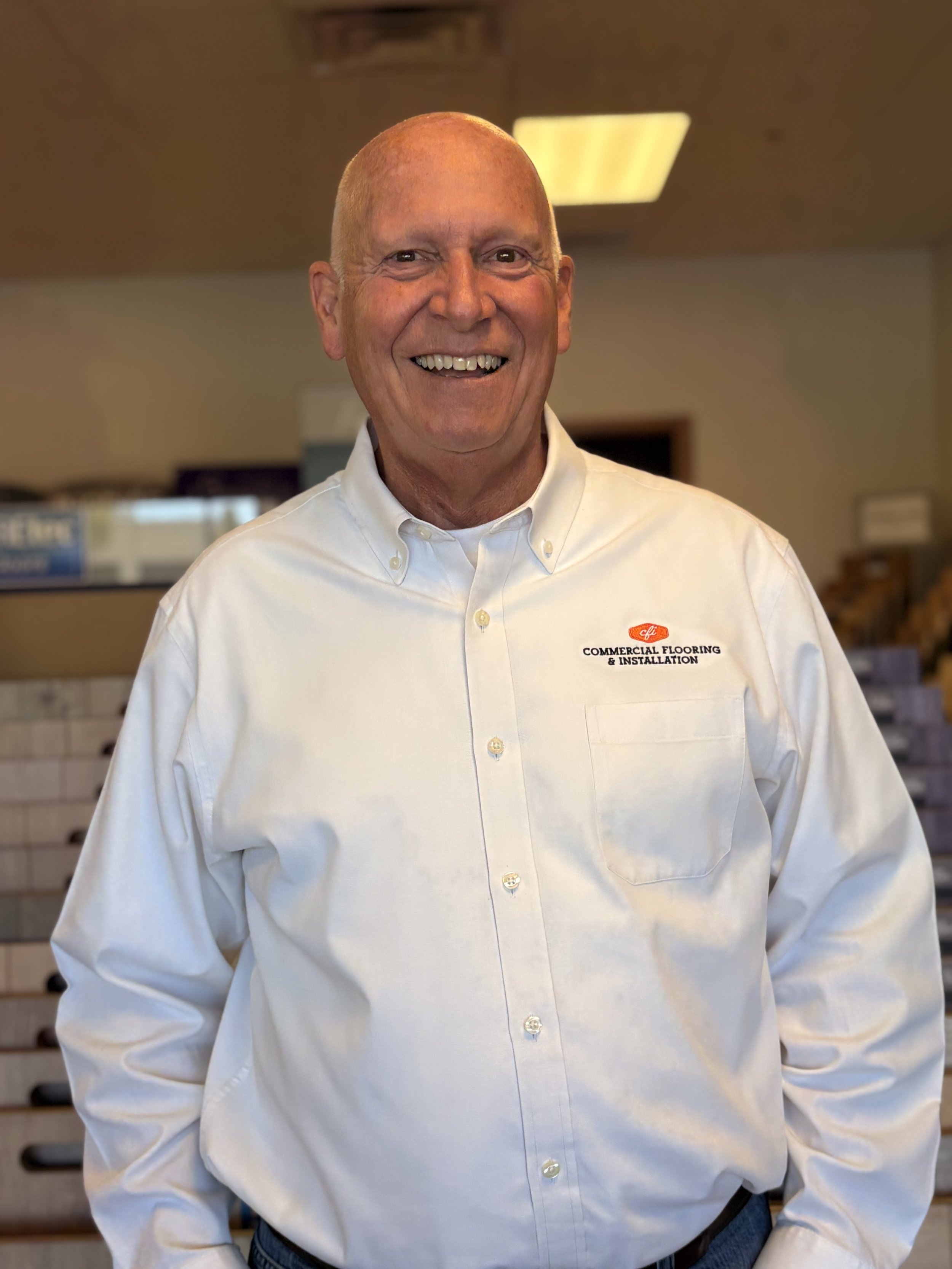 A smiling man in a white button-up shirt with a company logo, standing in front of a display of various tile samples in a flooring store.