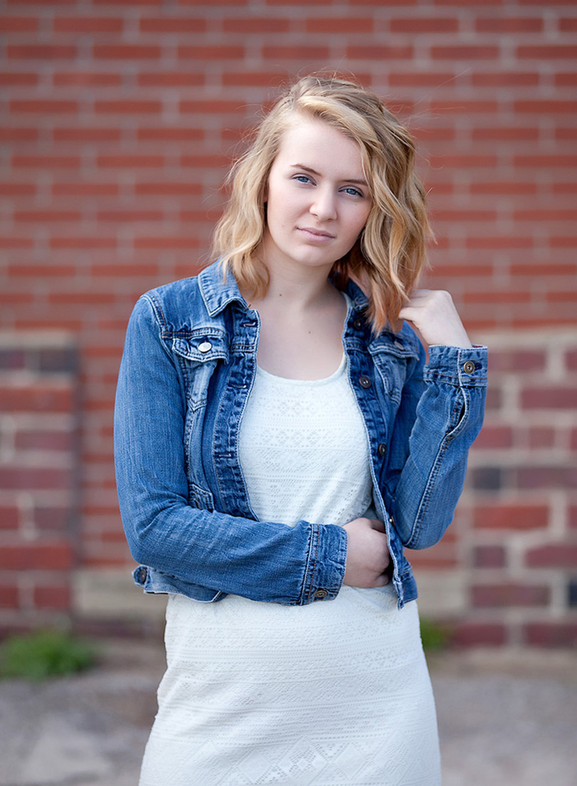 blonde senior girl with white dress and jean jacket in front of brick building in Orrville, Ohio