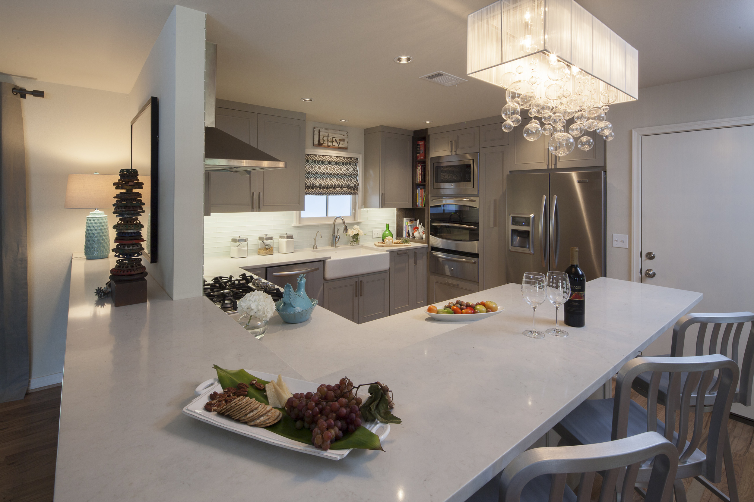Overview to the kitchen from the central area of the combination den+dining area. Notable is the large sweep of the counter-top which simultaneously serves multiple uses: Display surface for lamp and artwork; dining at the bar and food prep. Underneath and hidden in this view, are bookcase (at left) and the kitchen cabinetry with its " magic corners " and mixer lift . (Counter top material is caesarstone/quartz)