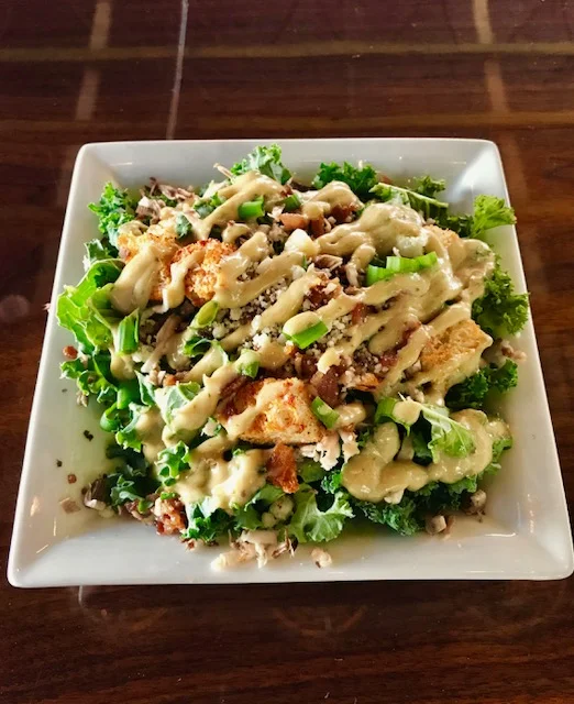 A square white plate filled with a salad topped with creamy dressing, chopped green onions, shredded cheese, croutons, and chopped nuts, placed on a dark wooden table.