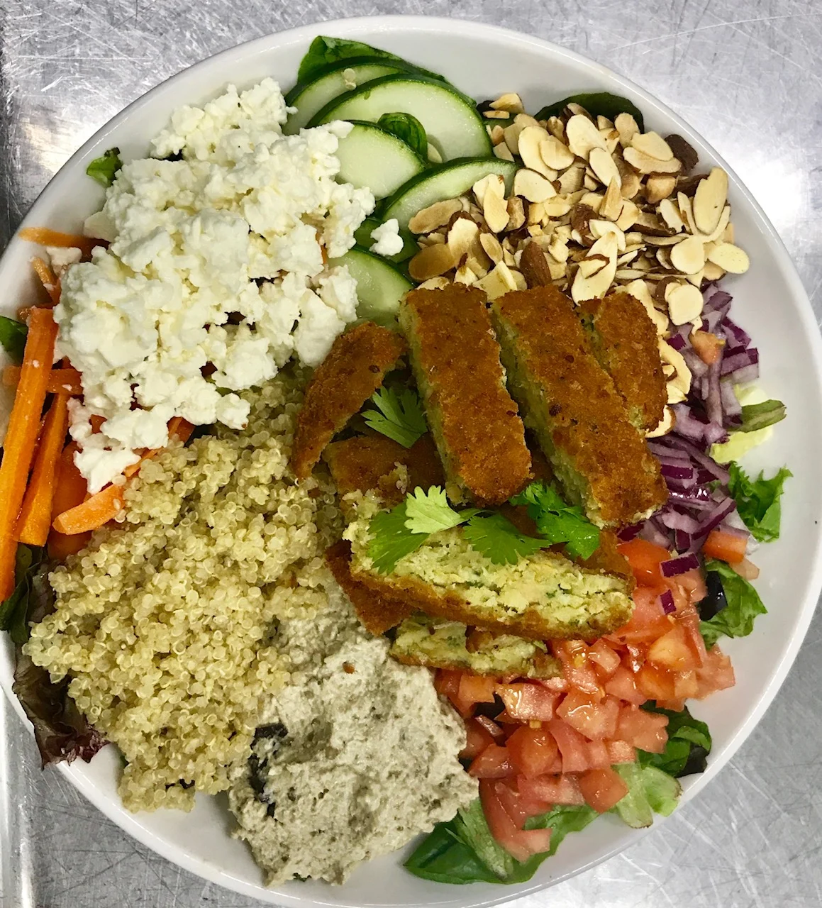 A white plate with a variety of vegetarian foods including falafel, chopped tomatoes, chopped onions, chopped carrots, cucumber slices, crumbled feta cheese, chopped nuts, couscous, and hummus.