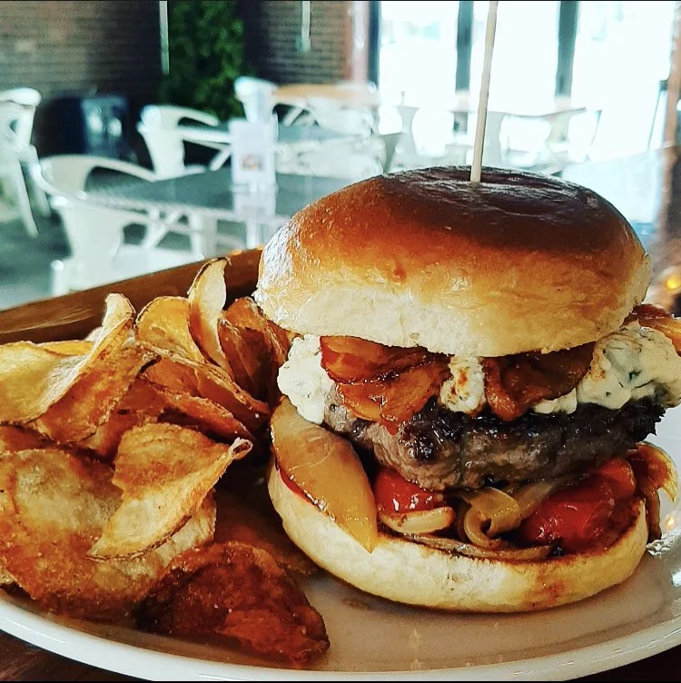 A cheeseburger with a beef patty, bacon, cheese, and vegetables in a bun, served with potato chips on a plate.