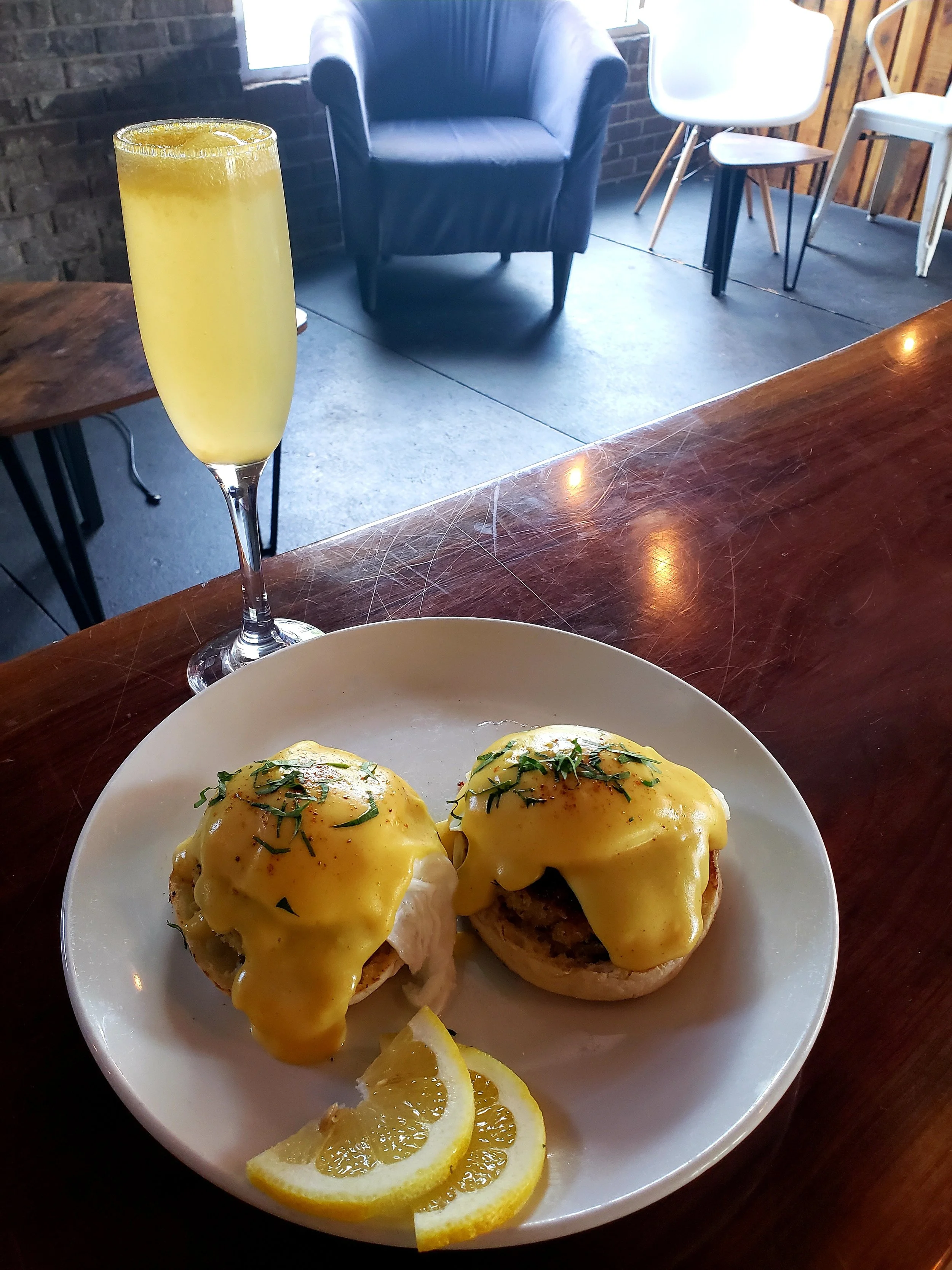 Two eggs Benedict with hollandaise sauce, garnished with herbs, served on a white plate with lemon slices, next to a glass of mimosa, on a wooden table.