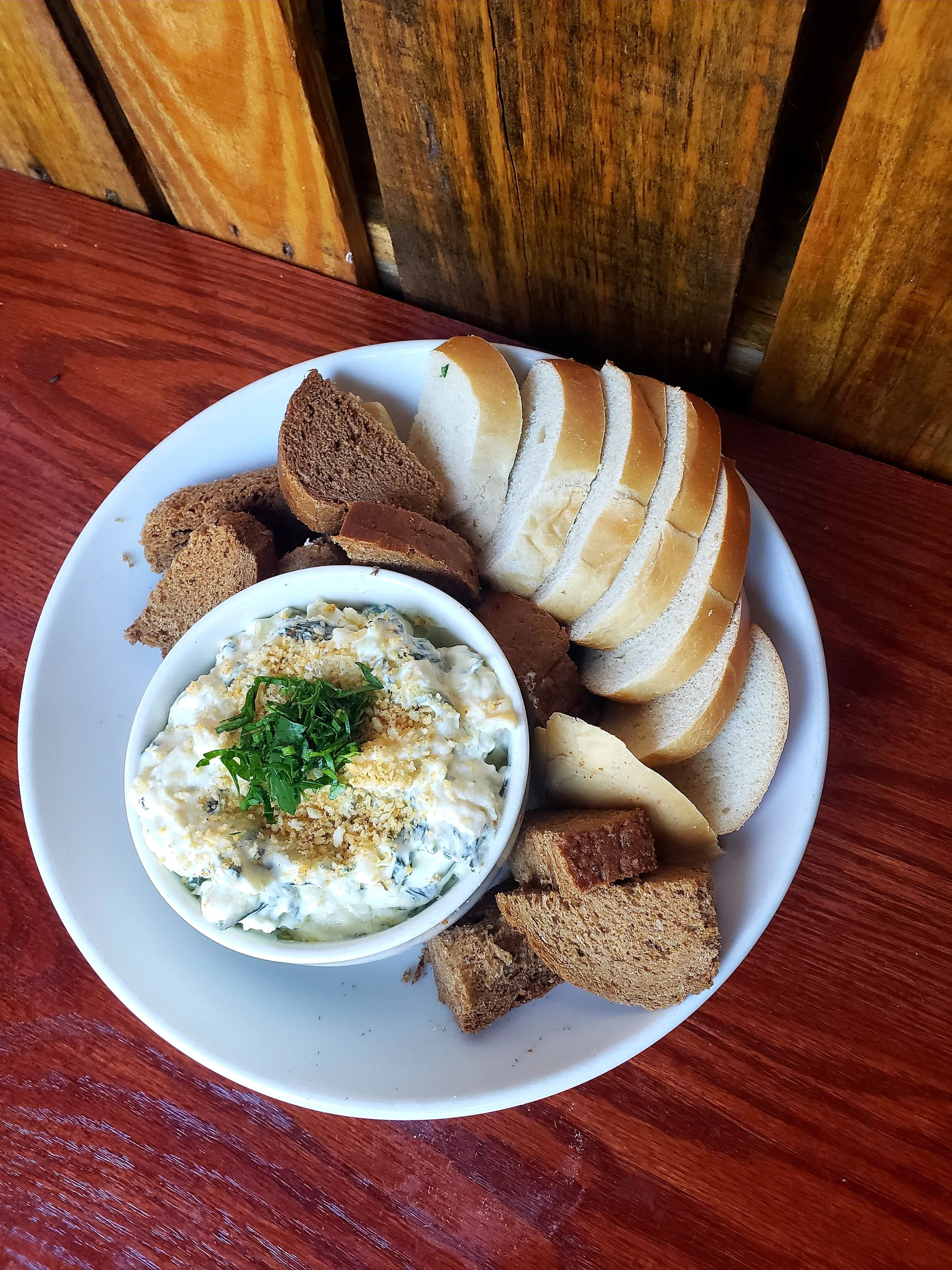 A white plate with sliced white bread, assorted brown bread, and a bowl of egg salad garnished with chopped herbs and crumbled crackers, all on a wooden table.