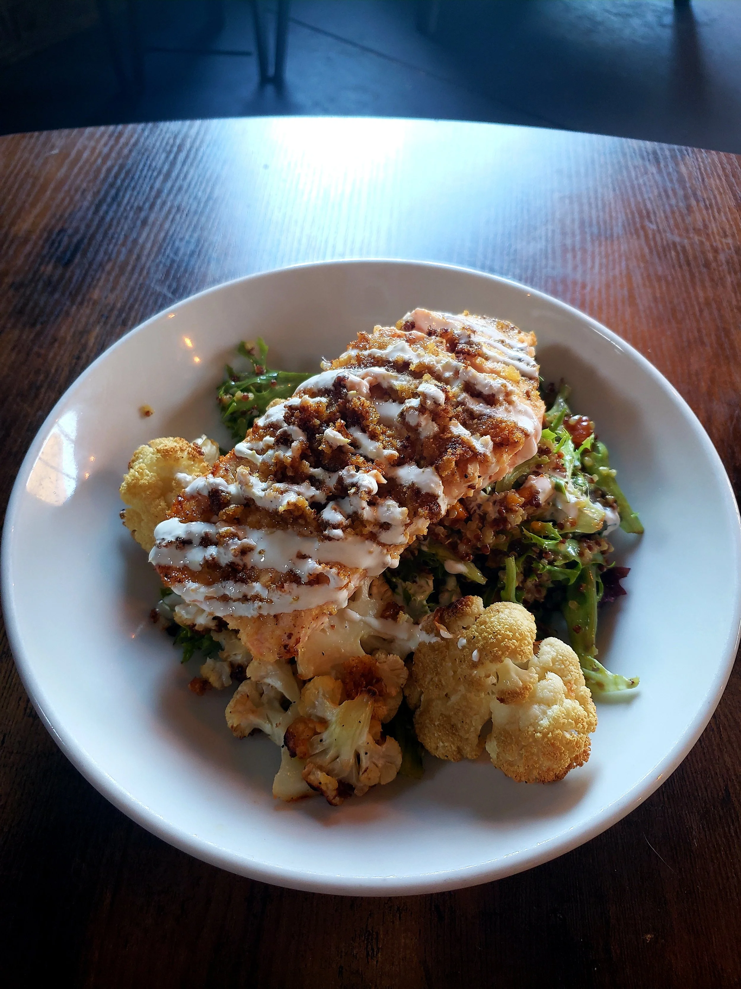 A white bowl contains a fried fish fillet topped with sauce and garnished with crumbled topping, served over a bed of mixed greens and roasted cauliflower on a wooden table.