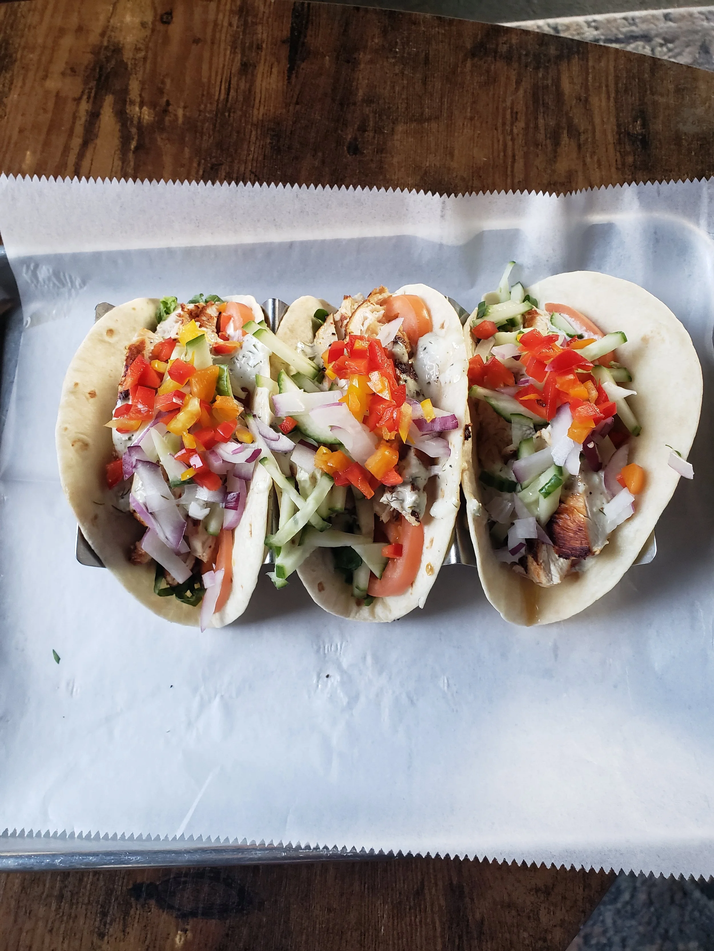 Three tacos filled with grilled meat, topped with chopped onions, tomatoes, and colorful peppers, served on a sheet of parchment paper.
