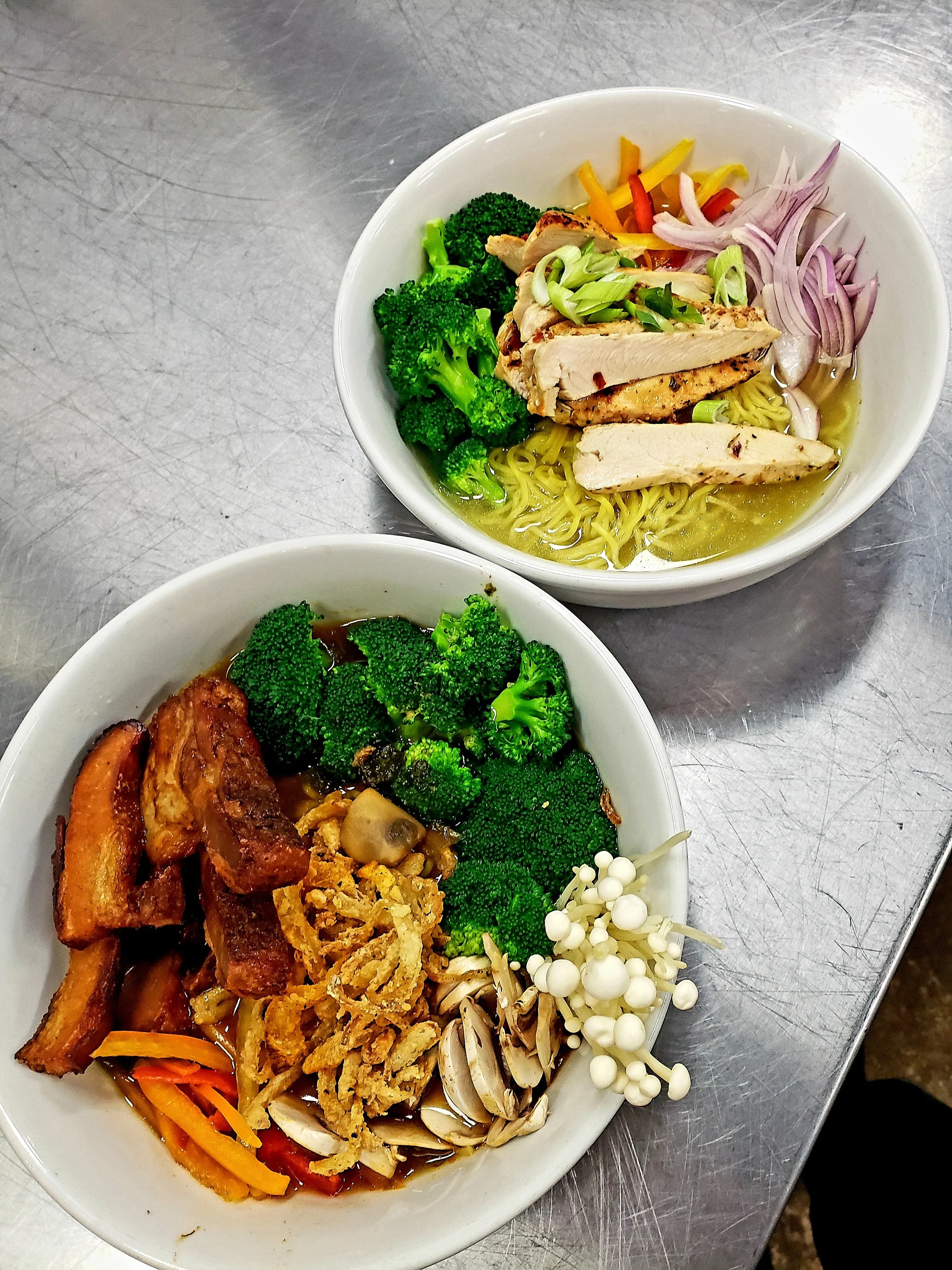 Two bowls of ramen with various vegetables and toppings on a metal surface.