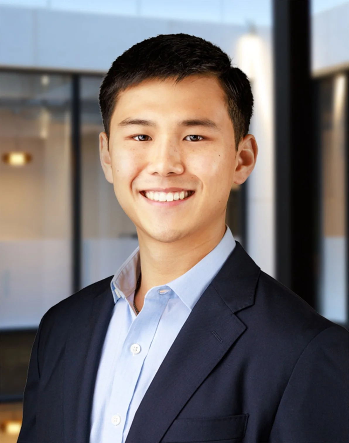 A young man wearing a dark suit and light blue collared shirt, smiling, standing in a modern office building.