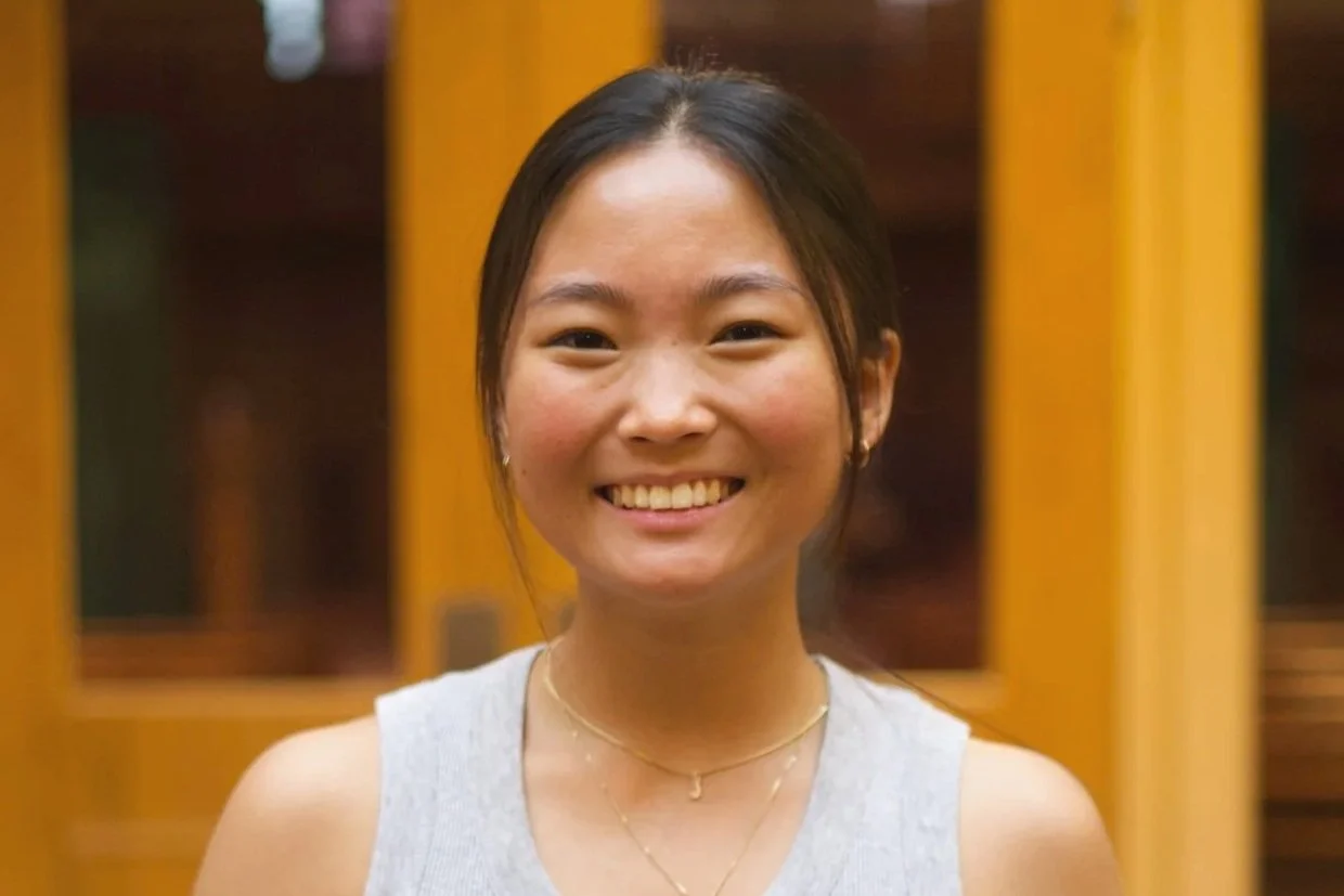 A young woman with dark hair smiling, wearing a sleeveless top and gold jewelry, standing indoors with wooden doors in the background.