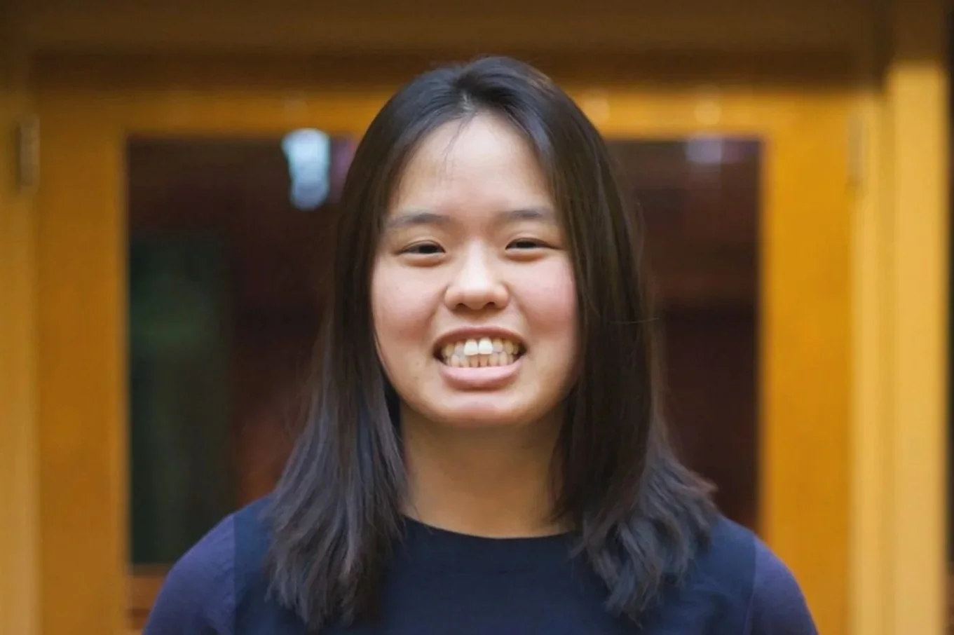 A young woman with shoulder-length dark hair, smiling with her teeth showing, standing indoors in front of a wooden structure.