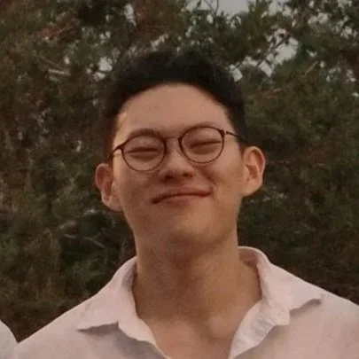 A young man with glasses and short dark hair, smiling outdoors with trees in the background.