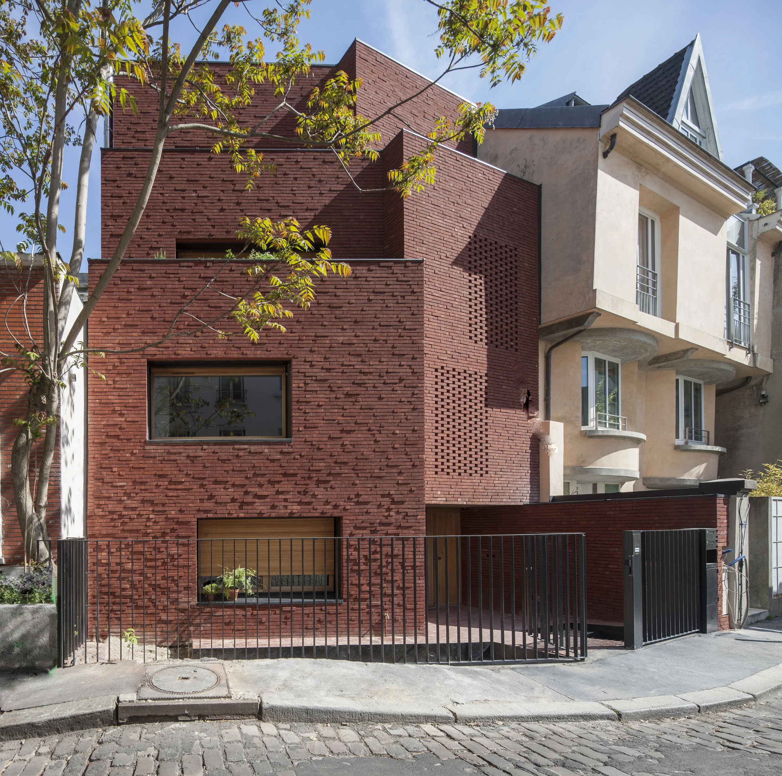 HOUSE ON THE BUTTE BERGEYRE The house is located in the Butte Bergeyre, a neighbourhood of houses and villas in northeast Paris, constructed on top of quarries and accessed by a single winding road. The plot of land sits on a bend in the road; the front façade of the house folds vertically, following the bend. Two volumes have been generated along each parallel side of the fold: a larger one containing interior living spaces, with expansive window bays; a smaller one containing stairs and bathrooms, with openings protected by moucharabié screening in brick. Interior spaces and natural light wrap around each other to create a continuing sense of openness on a highly compact site. Spaces fold and unfold as one moves through the house; only upon reaching the top floor is one afforded a clear view. Stepping back from the street as the facade rises, the successive balconies and roof terrace incorporate built-in planters, with the intention of becoming spectacularly overgrown after a few years, similar to the one-storey house covered in grapevines that had previously occupied the site. The brick pattern is irregular in width and depth, creating a relaxed, woven texture for the façade. Exterior doors, windows, and shutters are all in solid oak, with waterproofing elements in anthracite-colored zinc. Interior floors are covered in terrazzo and wide-plank oak parquet, consistent with the choice of natural, durable materials that comprise the entire construction. Bedroom partitioning has been created from oak storage units and sliding panels, giving flexibility to the floor layout as the family evolves. Project featured in “AMC 100 Bâtiments de l’année 2018”. La Butte Bergeyre est un quartier idyllique de maisons et de petits immeubles, à l'abri de la clameur du Nord-Est parisien et accessible par une seule route sinueuse. En contraste direct avec l'uniformité du Paris Haussmannien, le quartier conserve un air villageois éclectique grâce à des façades variées et à une végétation abondante, dont un vignoble en coteau qui marque l'entrée du quartier. Les clients de la maison, une jeune famille de quatre personnes, ont exigé une construction durable qui permet à la végétation de l'entourer au fil du temps. Une banale maison de 45 m2 de plain-pied a été remplacée par un petit building de quatre étages. La position de la parcelle dans une courbe de la rue a engendré la forme à facettes du bâtiment, la façade sur rue se repliant verticalement pour épouser le virage. Deux volumes ont été créés de chaque côté du pli : le plus grand contient les espaces de vie, avec de spacieuses baies vitrées, et le plus petit enferme les escaliers et les salles de bains, l’ensemble bénéficiant d’ouvertures abritées par des moucharabieh en brique. Les espaces intérieurs et la lumière naturelle s'enroulent l’un sur l’autre pour émuler une sensation permanente d’ouverture sur ce site très compact. Les espaces se déploient au fur et à mesure que l'on se déplace et que l'on monte aux étages ; en atteignant l'étage supérieur, on profite d’une vue parfaitement dégagée. Depuis la rue, au fur et à mesure que l’on s’éloigne de la façade, on découvre les jardinières encastrées dans les balcons successifs, dont les plantations sont destinées à croître spectaculairement après quelques années et à taquiner à nouveau les vieilles vignes de la façade. Le motif de brique joue sur des largeurs et des profondeurs irrégulières pour créer une texture de façade tissée et décontractée. Les portes, les fenêtres et les volets extérieurs sont en chêne massif et des éléments d'étanchéité en zinc anthracite. Les sols intérieurs sont recouverts de granito et de parquet en chêne à lames larges, ce qui correspond au choix de matériaux naturels et durables qui se patineront au fil du temps. Il y a peu de murs intérieurs. Les cloisons des chambres à coucher sont composées d'éléments de rangement et de panneaux coulissants en chêne, ce qui donne de la souplesse à l'aménagement des espaces en fonction des besoins d’une famille évolutive. https://www.amc-archi.com/photos/lia-kiladis-maison-et-studio-paris,8164/lia-kiladis-maison-et-studio.1
