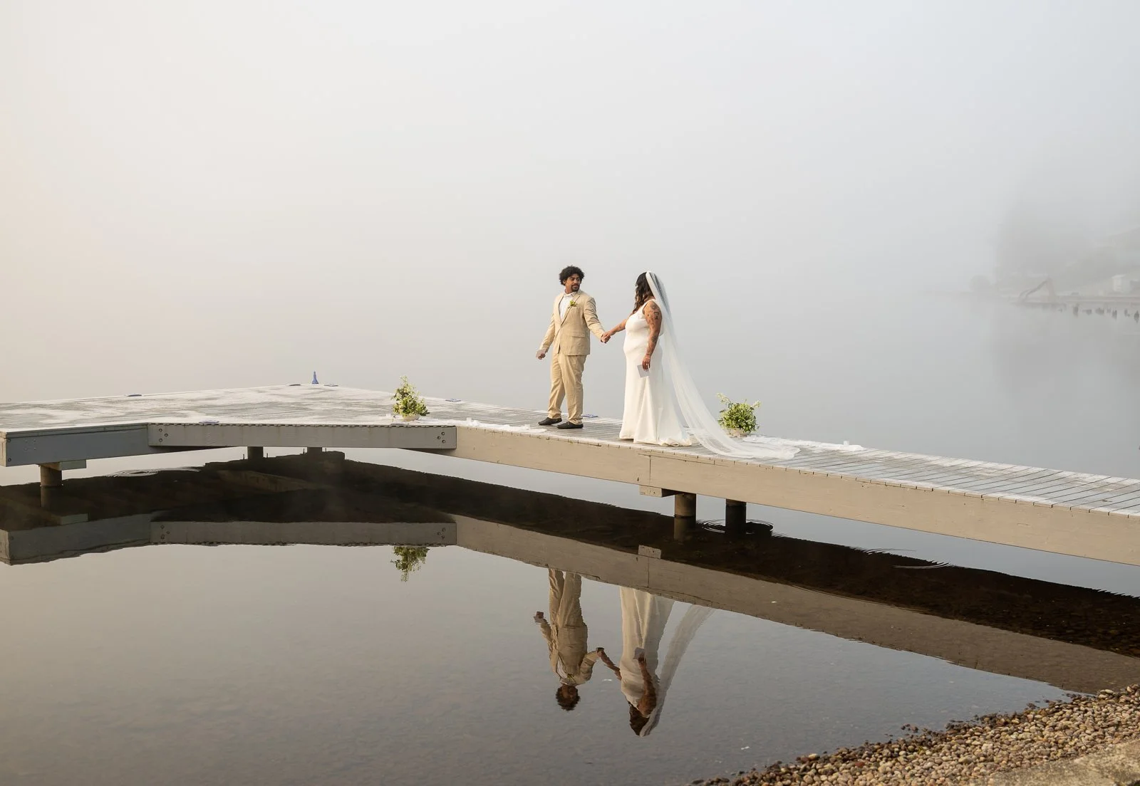 Groom glancing back at the bride as they walk down the dock, her veil trailing behind them through the fog and marina.