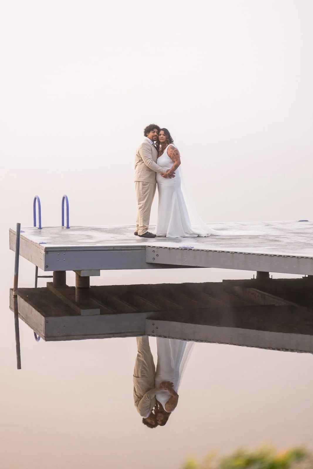 Bride and groom embracing at the end of the dock as the fog turns soft pink at sunrise, their reflection glowing in the still water below.