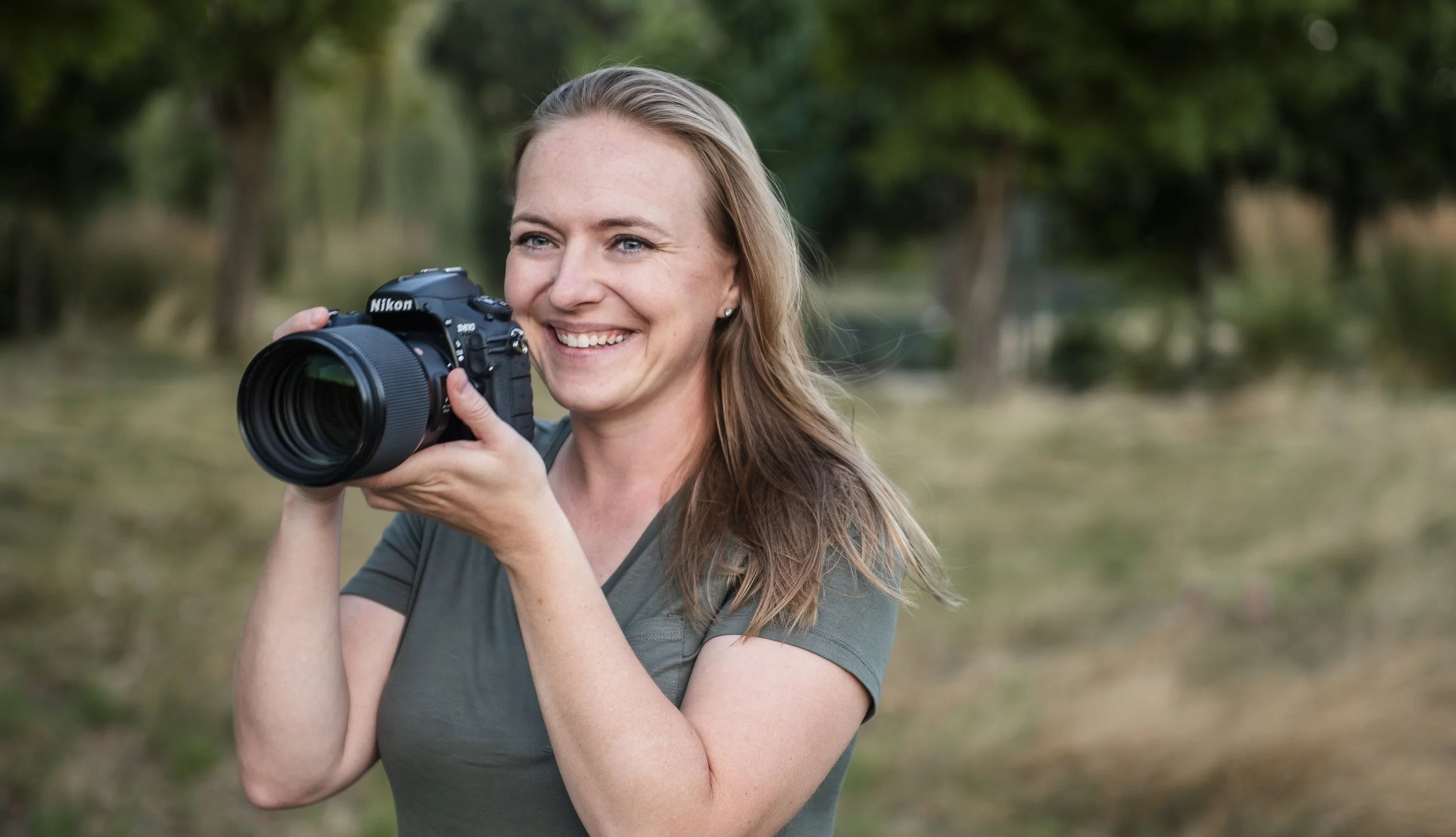 A woman in a green shirt, outside, holding a camera up to her face with a smile, and a twinkle in her eye