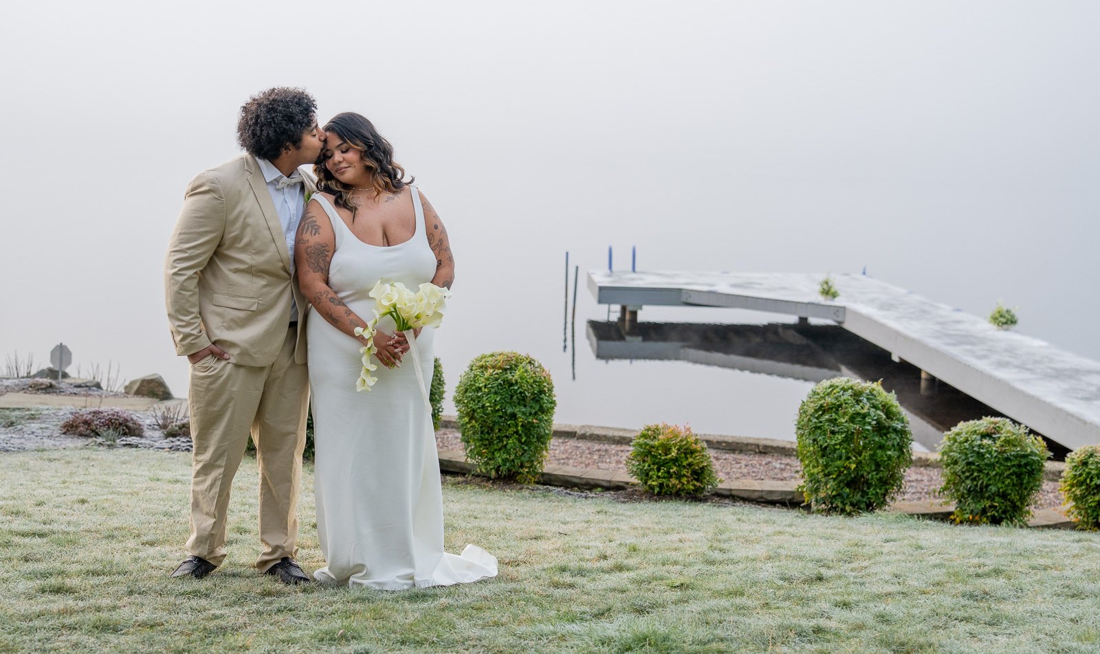 Groom kissing the bride's forehead as they embrace on the lawn, the dock and foggy lake visible behind them among the bushes.