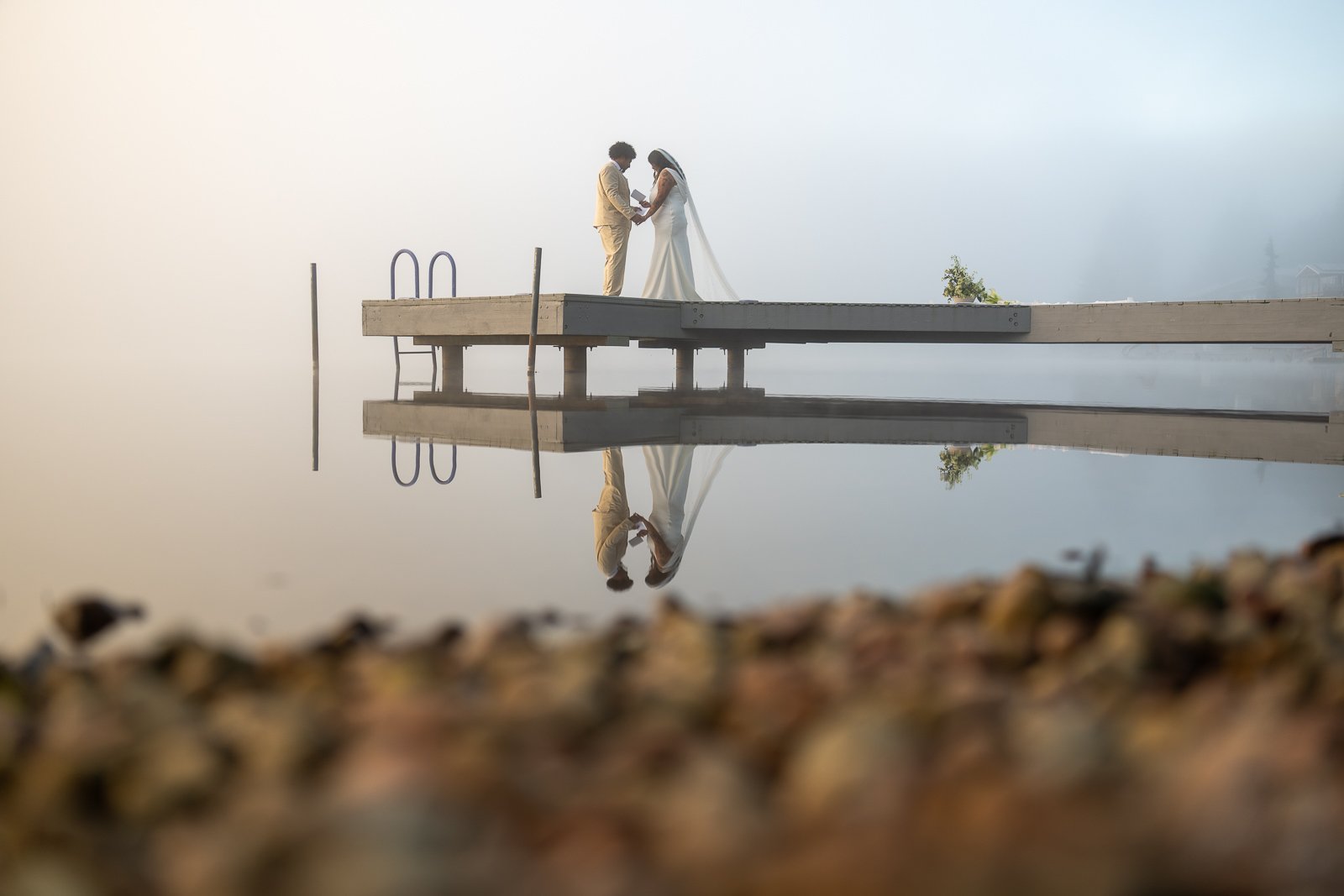 Bride and groom exchanging vows at the end of the dock, their reflection mirrored in the still lake water, shot through blurred foreground stones in Olympia, Wa