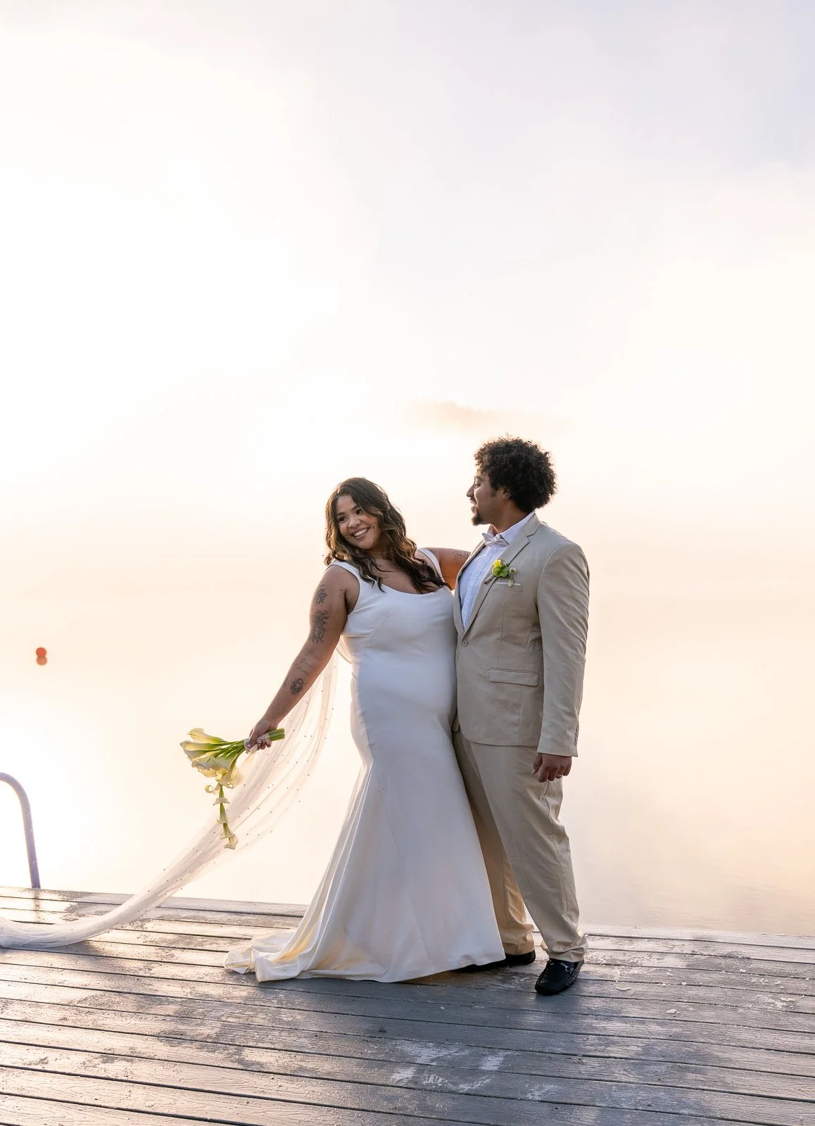 Bride and groom smiling at each other on the dock, her calla lily bouquet and veil catching the breeze in the fog.