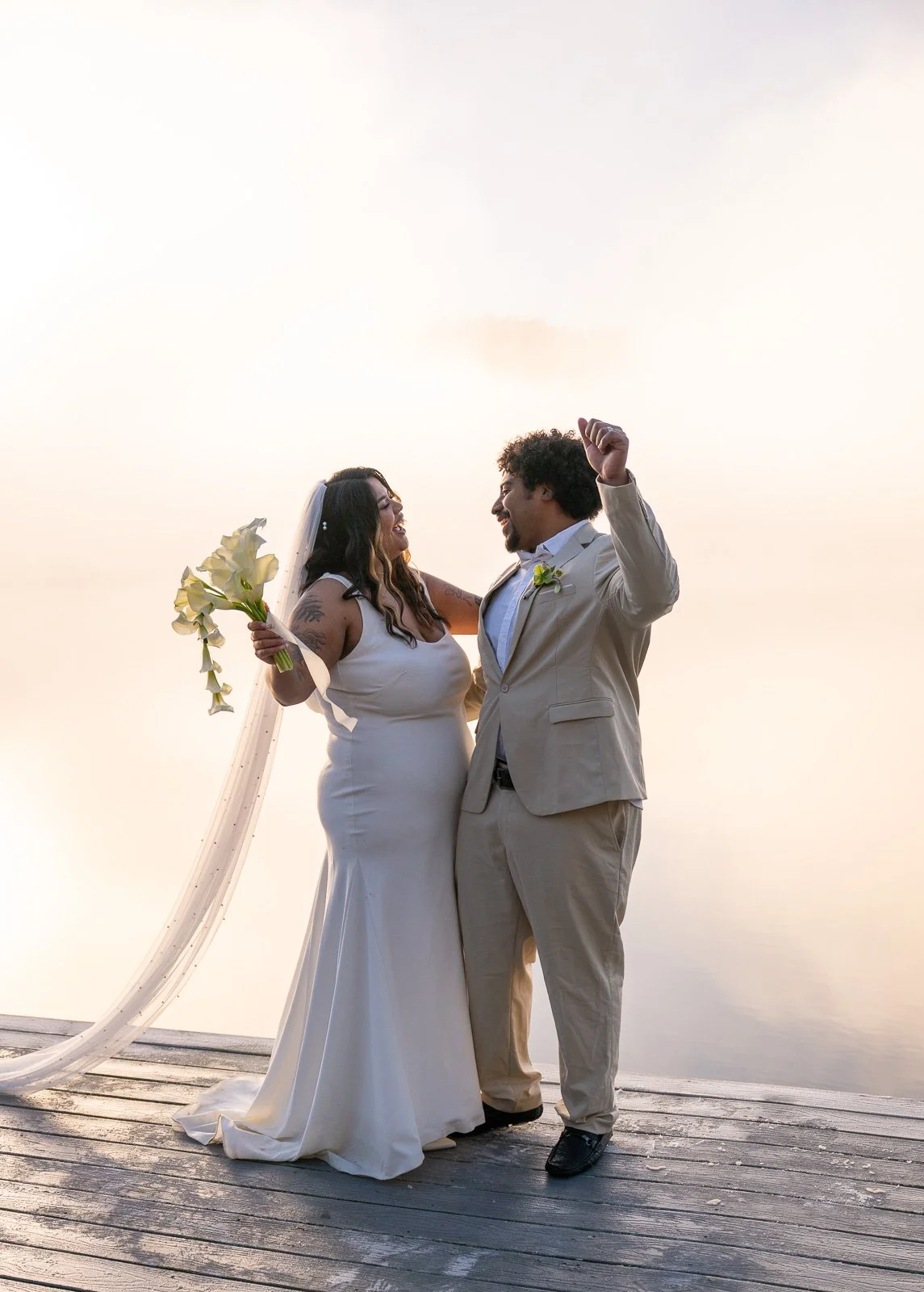 Bride and groom laughing together on the dock, groom's hand raised mid-twirl, morning fog rising off the lake behind them.