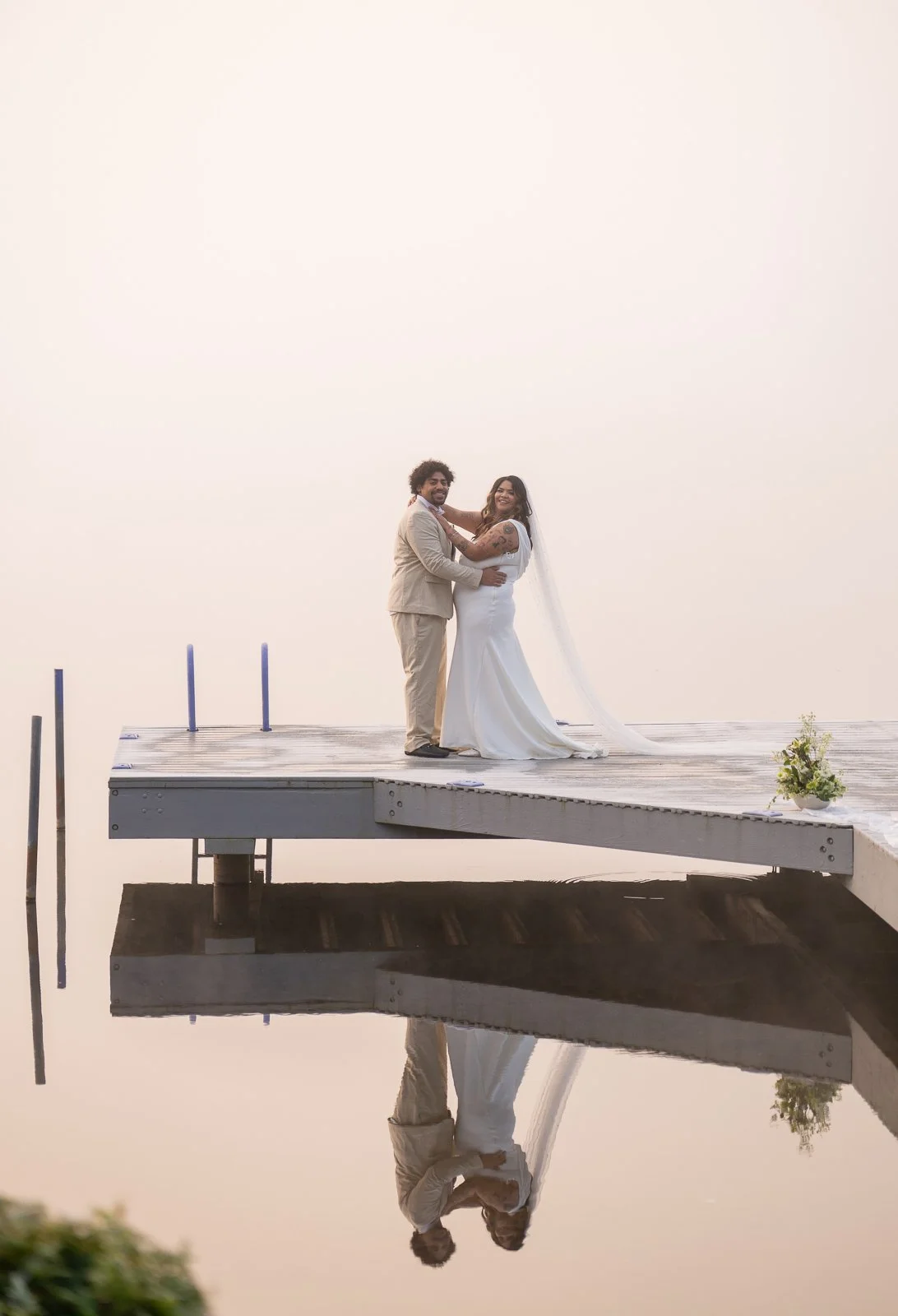 Bride and groom standing close together at the end of the dock, embracing and looking toward the camera, their reflection visible below.