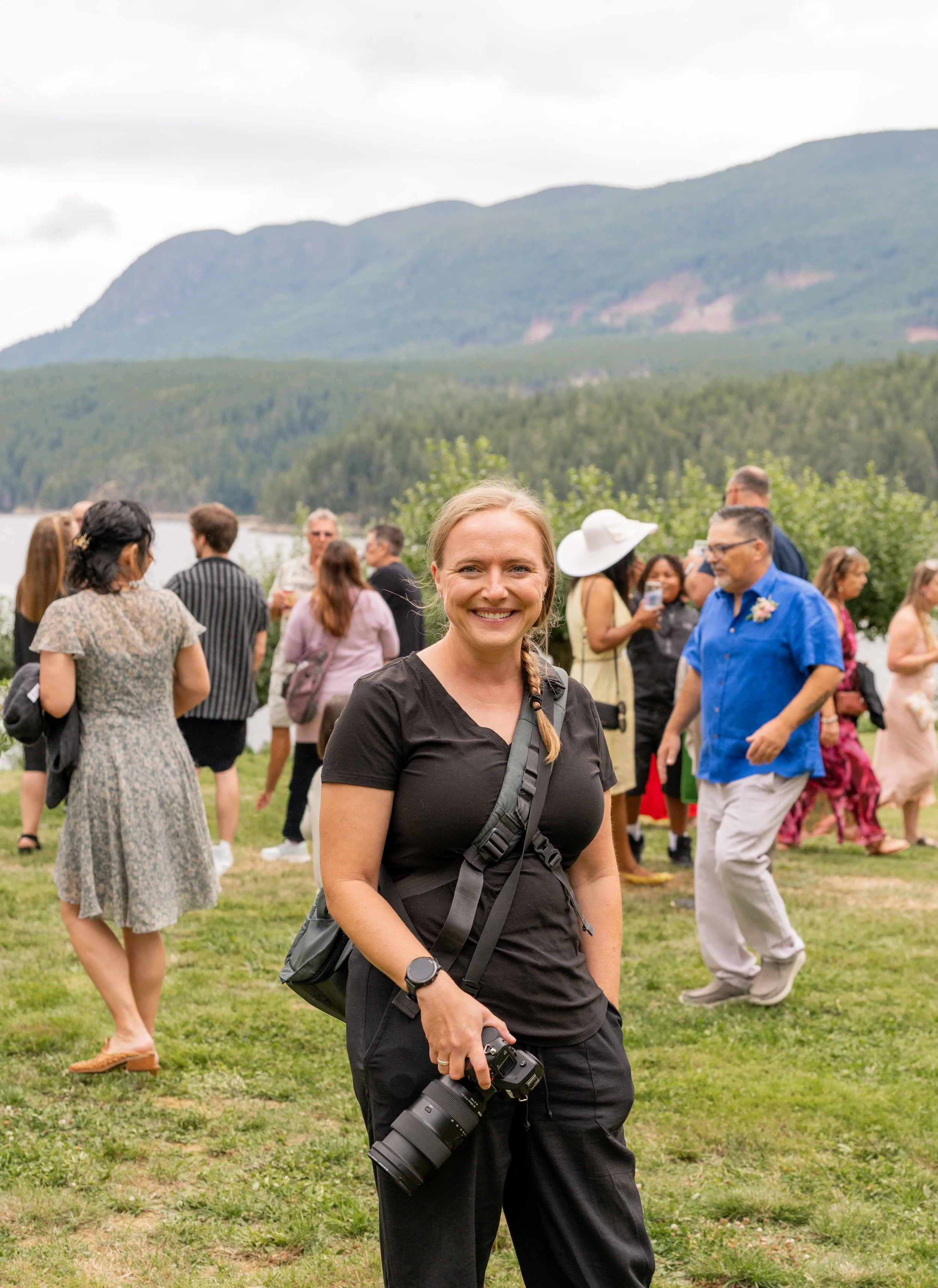 A woman dressed in all black, holding her camera, at a wedding, with a mountain and the ocean in the background