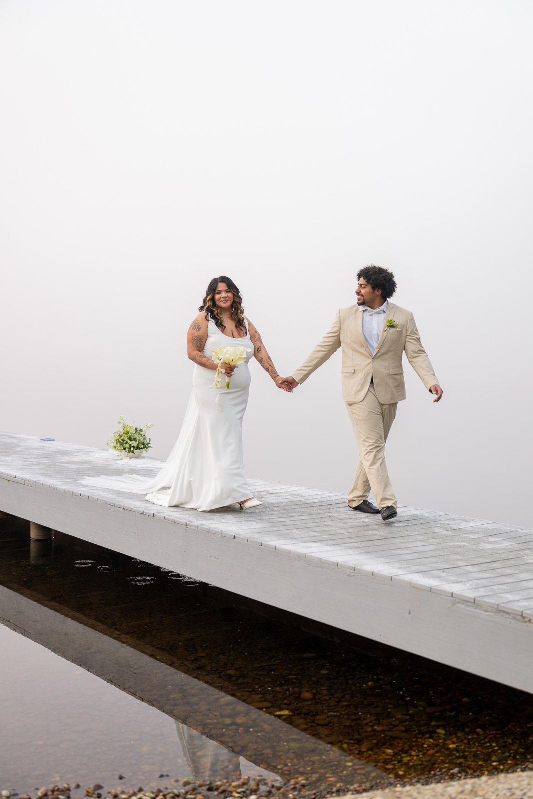 Bride and groom walking hand in hand down the dock past floral arrangements, smiling at each other in the morning fog.